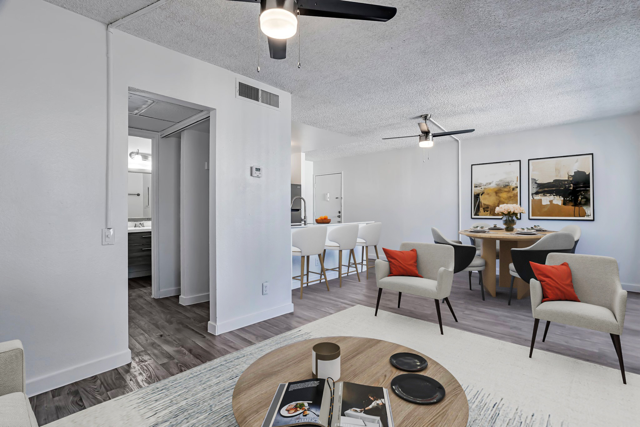 A modern, brightly lit living space featuring a round dining table with chairs, two armchairs, and a coffee table. The decor includes art prints on the wall, a cozy rug, and a ceiling fan. The open layout connects to a kitchen area visible in the background.