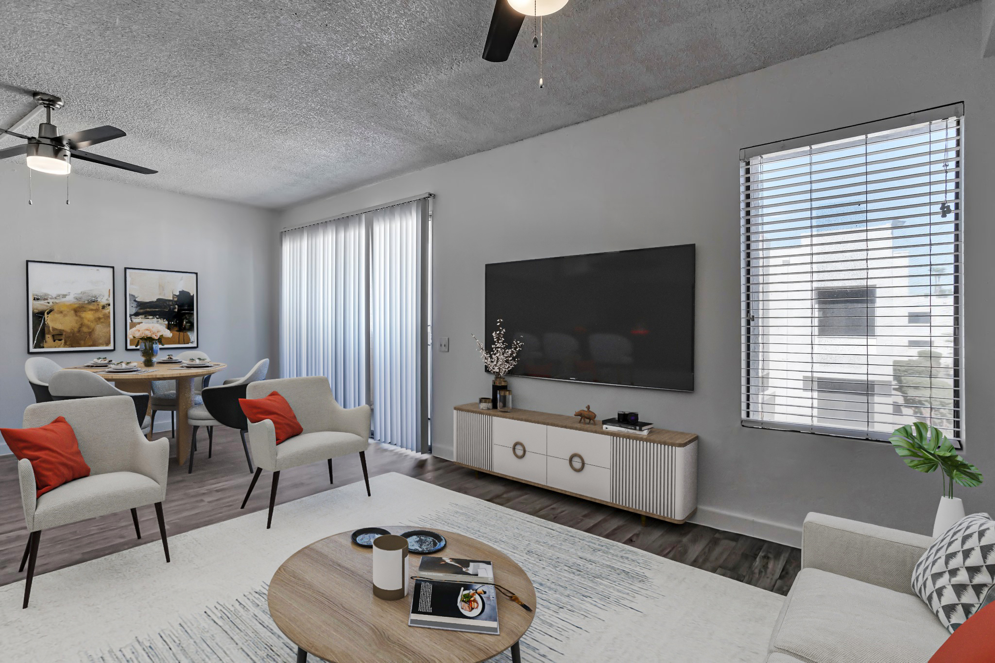 A modern living room featuring two ceiling fans, a large flat-screen TV mounted on a wall, and a stylish media console. There are two comfortable armchairs with red accent pillows and a coffee table with decorative items. Bright natural light comes in through large windows with blinds, complementing the minimalist decor.