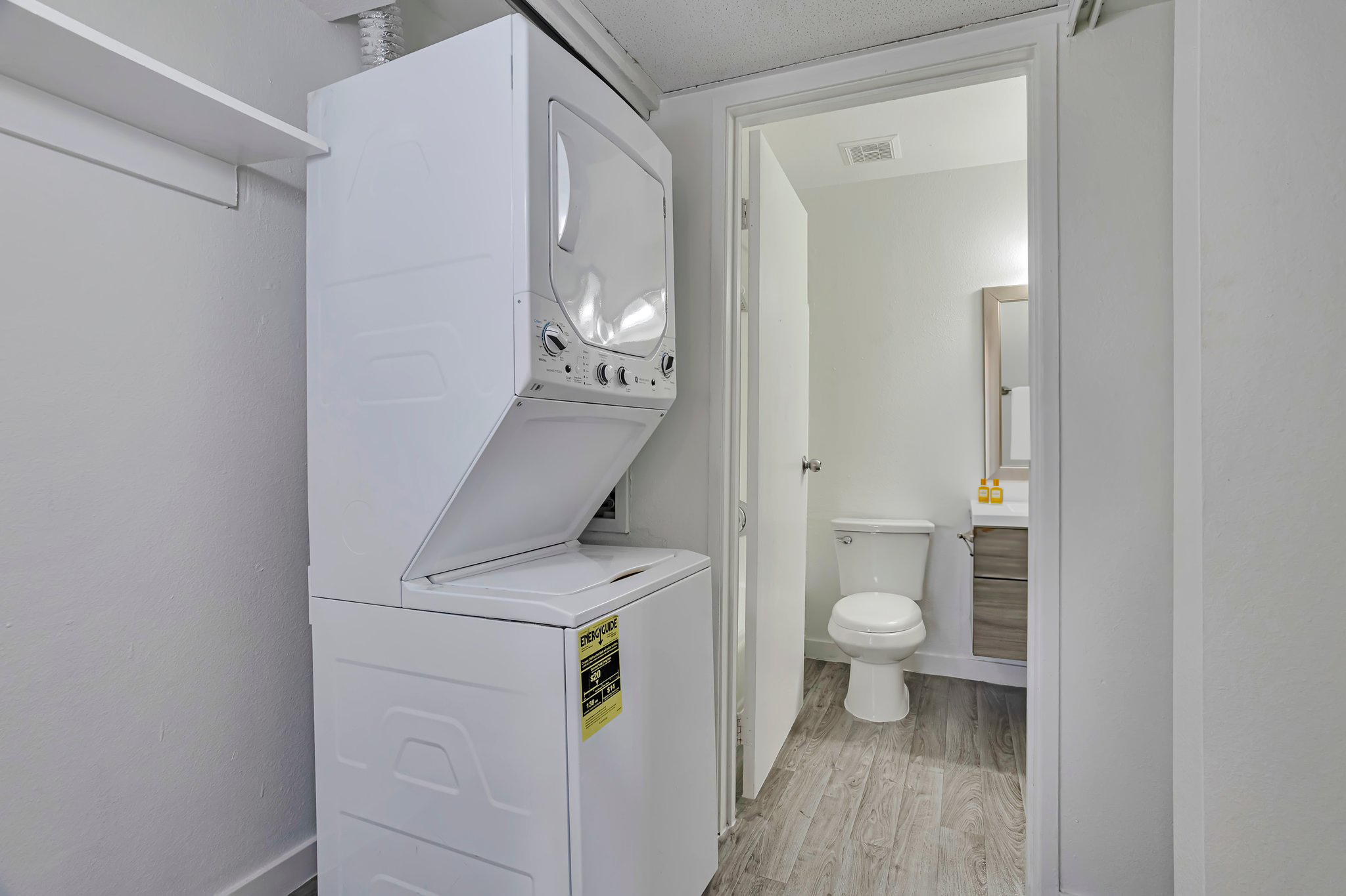 A laundry area featuring a stacked washer and dryer against a wall, with a doorway leading to a bathroom. The bathroom includes a toilet, a vanity with a mirror, and white walls. Light-colored flooring enhances the bright, clean look of the space.