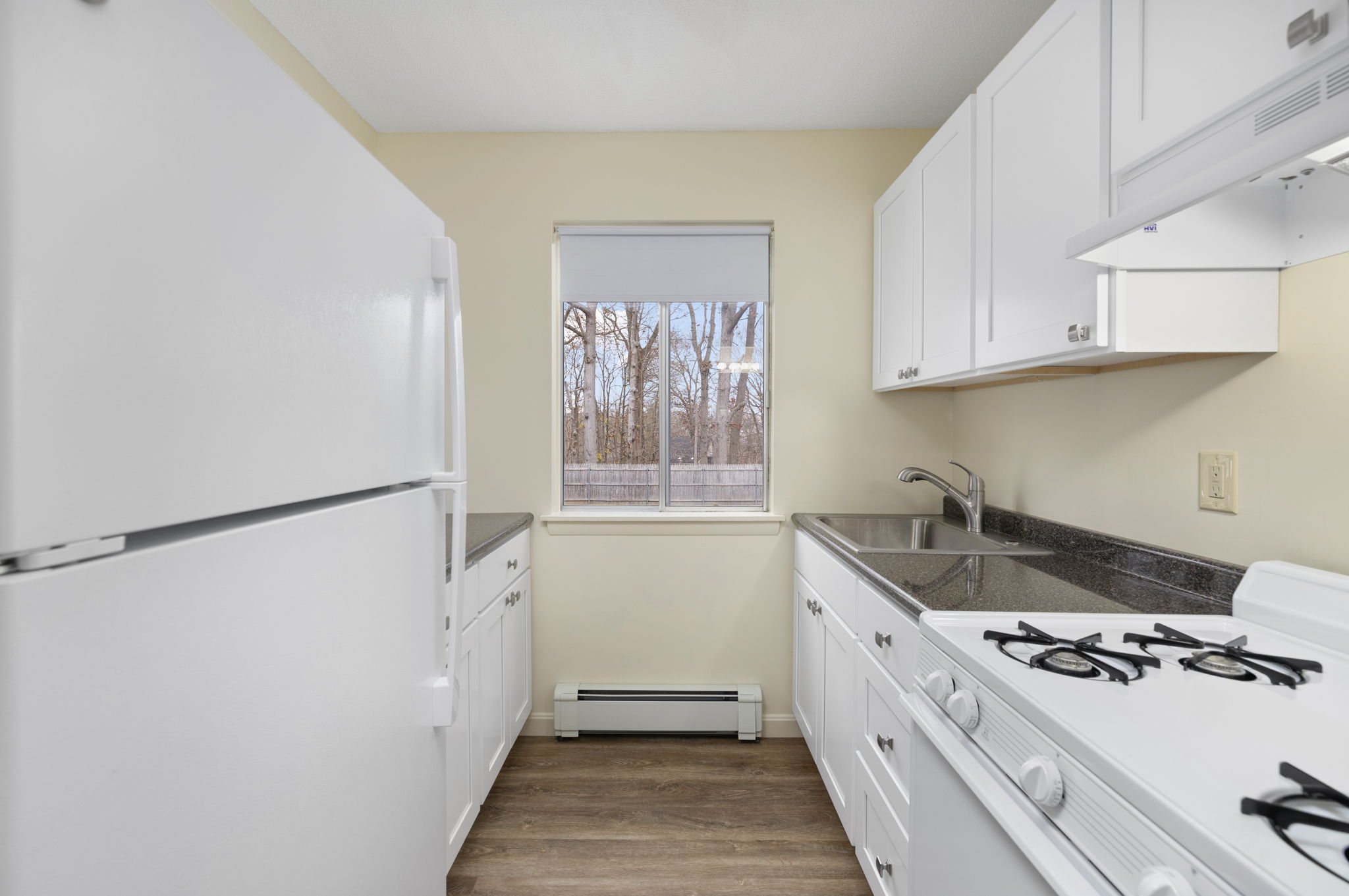 Bright kitchen featuring white cabinets, a large white refrigerator, and a gas stove. A sink is positioned under a window that allows natural light in, showcasing a view of trees outside. The flooring is wood-like, and the overall decor is clean and modern.