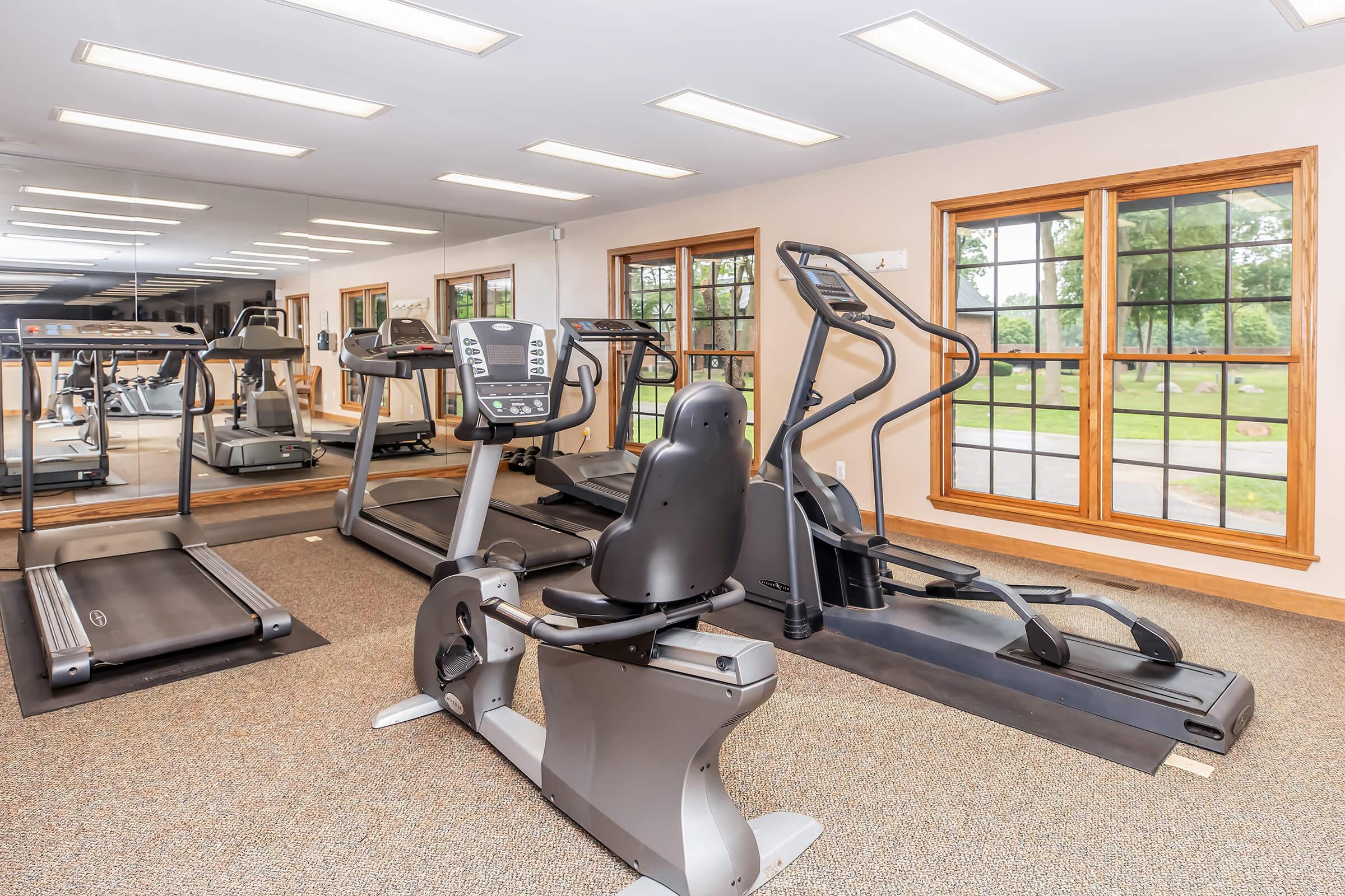 A fitness area featuring a treadmill, an elliptical machine, and a stationary bike, all positioned against mirrored walls. Natural light filters in through large windows, enhancing the space's inviting atmosphere. The floor is carpeted, and the decor is simple, promoting a clean and modern gym environment.