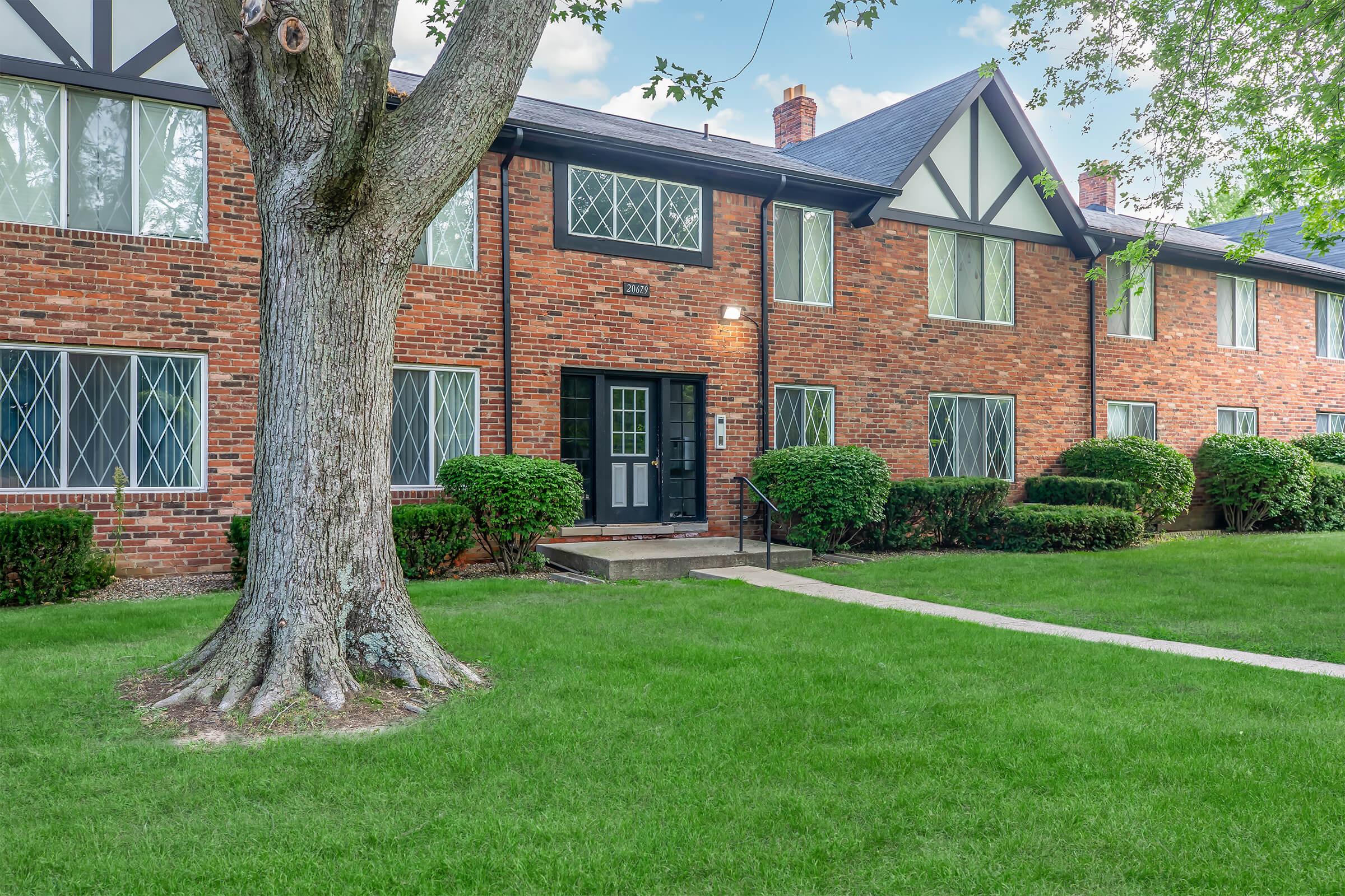 A brick apartment building featuring multiple units with large windows, surrounded by well-maintained green lawns and shrubbery. A mature tree stands in the foreground, providing shade, while a pathway leads to the entrance. The overall setting is neat and inviting.