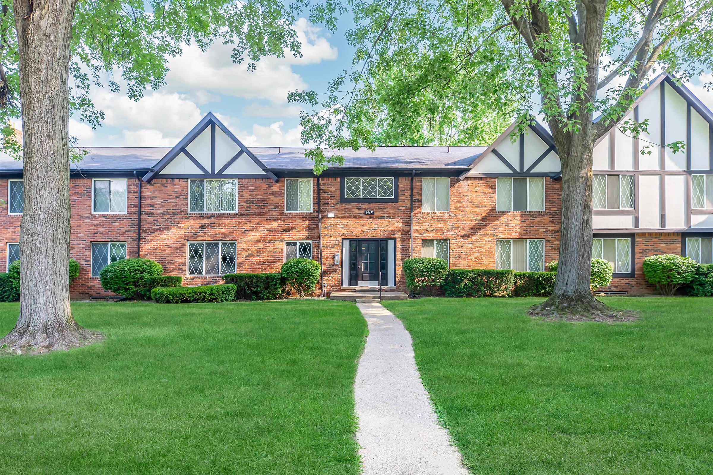 Brick apartment building with large windows, surrounded by manicured lawns and trees. A concrete pathway leads to the entrance, creating a welcoming atmosphere. The sky is bright with fluffy clouds, contributing to a peaceful, suburban setting.