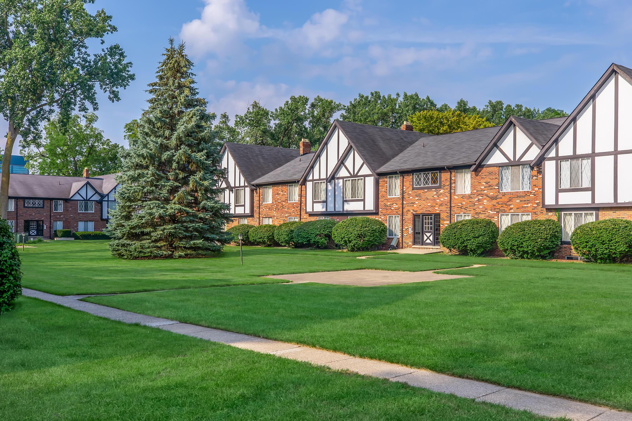 View of a well-maintained residential area featuring brick apartment buildings with Tudor-style architecture. The buildings are surrounded by lush green lawns and neatly trimmed bushes, with a paved walkway leading through the grass. Tall trees are visible in the background under a bright blue sky with scattered clouds.