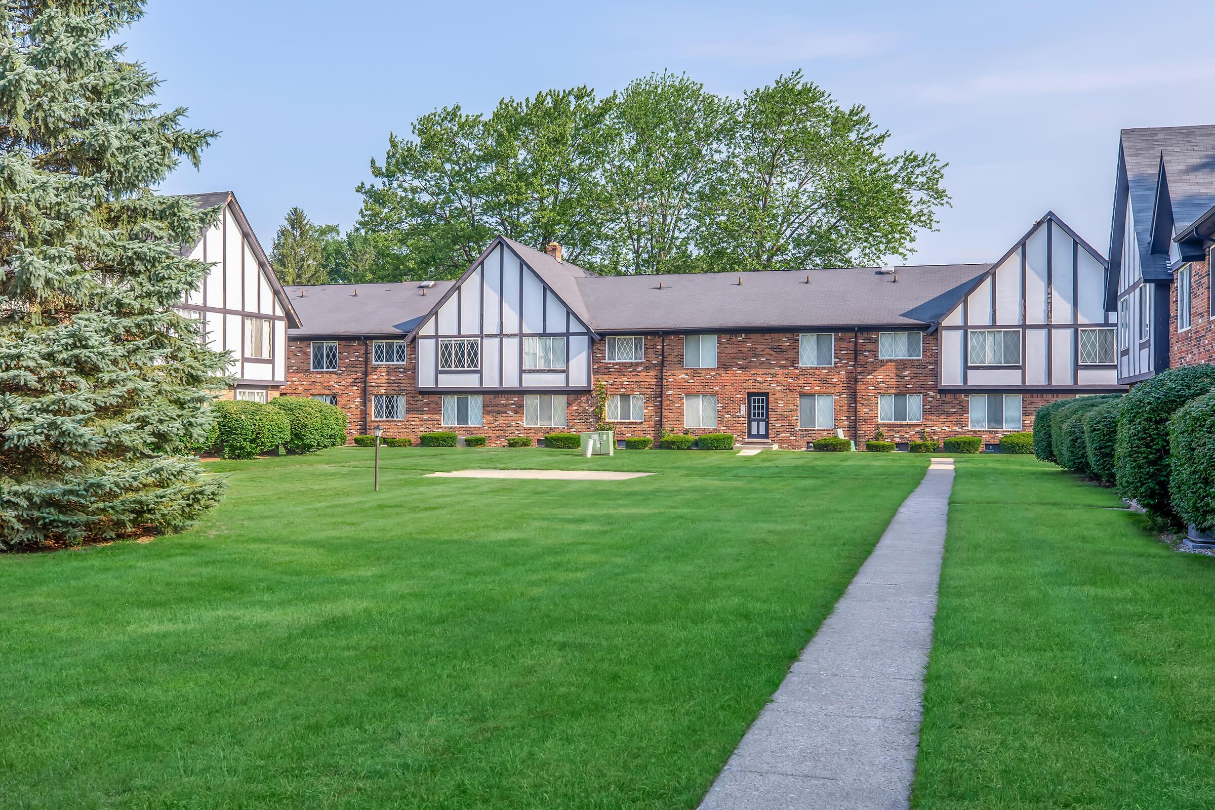 A well-maintained brick apartment building featuring a landscaped lawn with neatly trimmed grass and bushes. The structure displays a classic Tudor style, with multiple windows and gabled roofs. A paved walkway leads through the lush green area, connecting the building to the surrounding landscape.