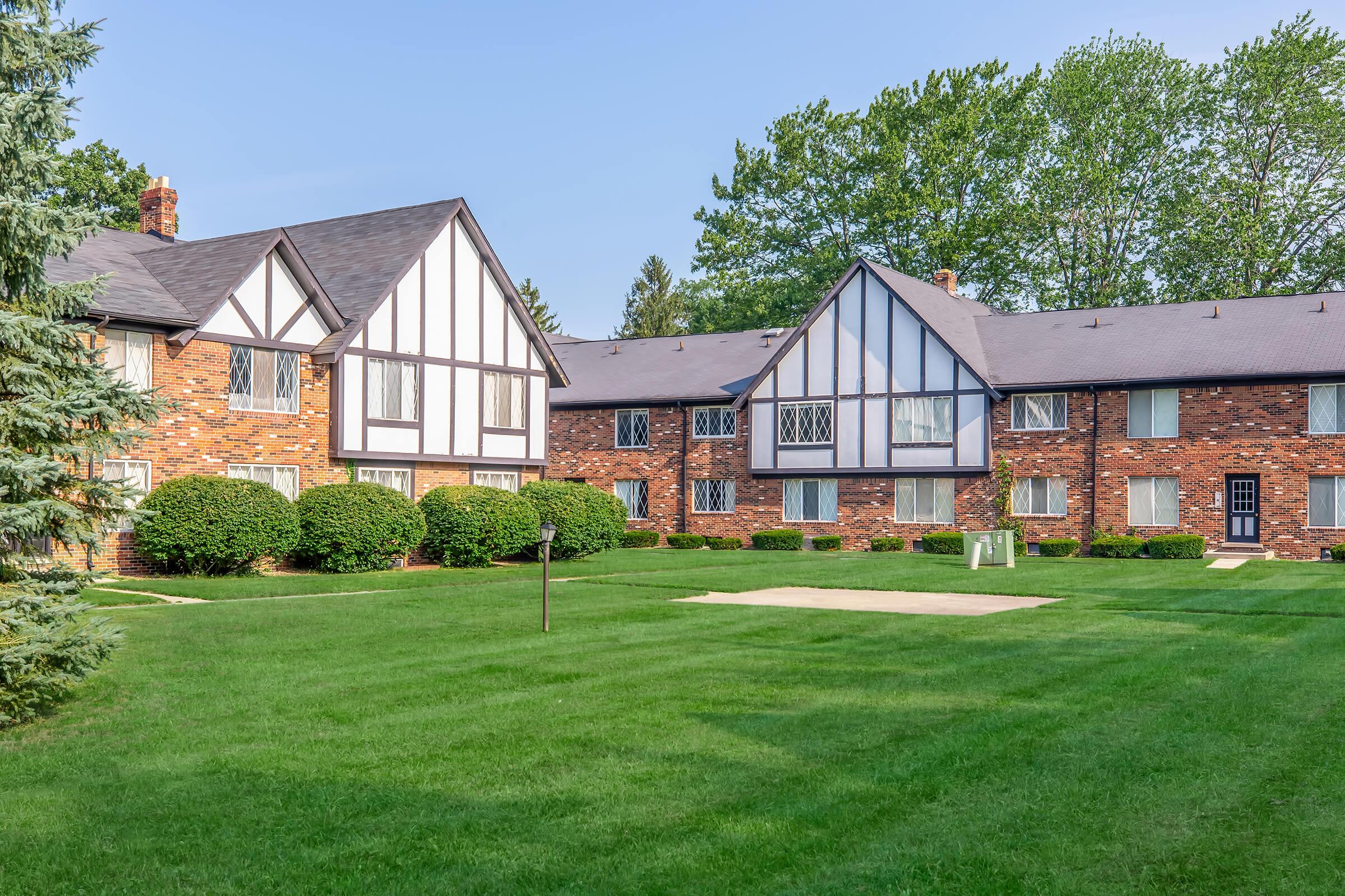 Two multi-story brick buildings with a Tudor-style design are surrounded by well-maintained green grass and shrubs. There’s a sandy area in front, indicating a possible recreational space. The sky is clear, and trees line the background, creating a serene setting.