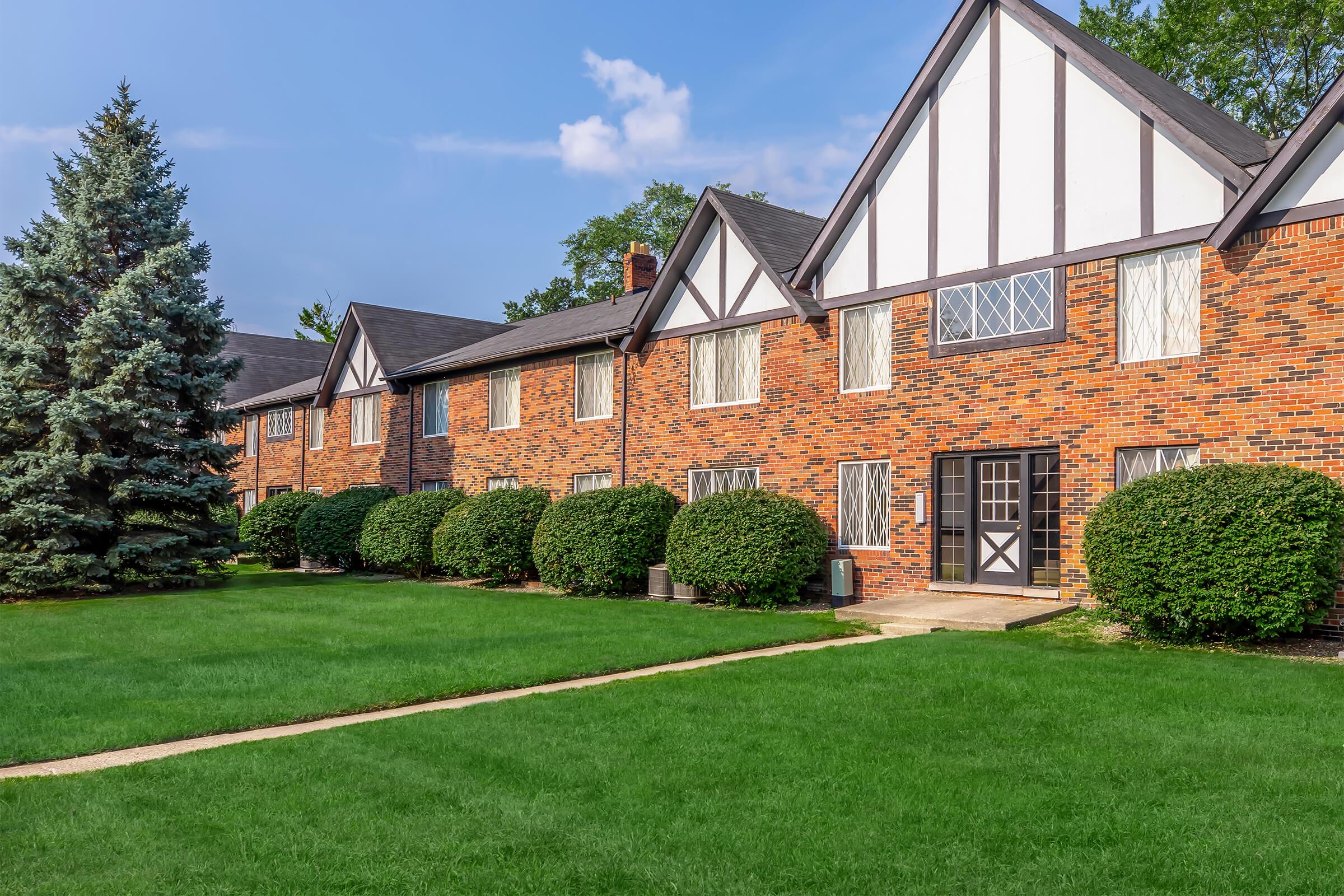 A brick apartment building with Tudor-style architecture featuring gabled roofs. The façade includes multiple windows with white frames. Lush green grass surrounds the building, and neatly trimmed bushes line the pathway leading to the entrance. Tall evergreen trees are visible in the background under a blue sky.