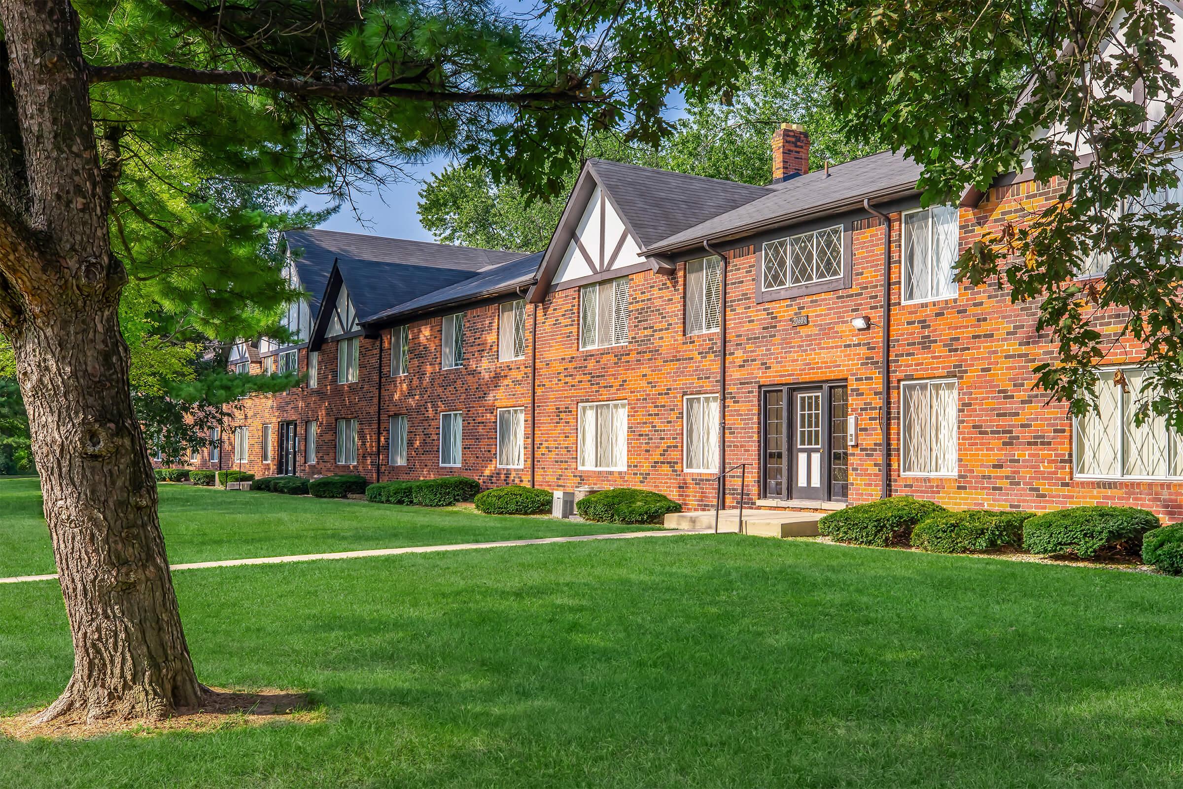 Brick apartment building with multiple units, featuring large windows and manicured lawns. The exterior is bordered by several trees, creating a pleasant, green space. Pathways connect the building to the surrounding grass areas. Clear blue sky above adds to the inviting atmosphere.