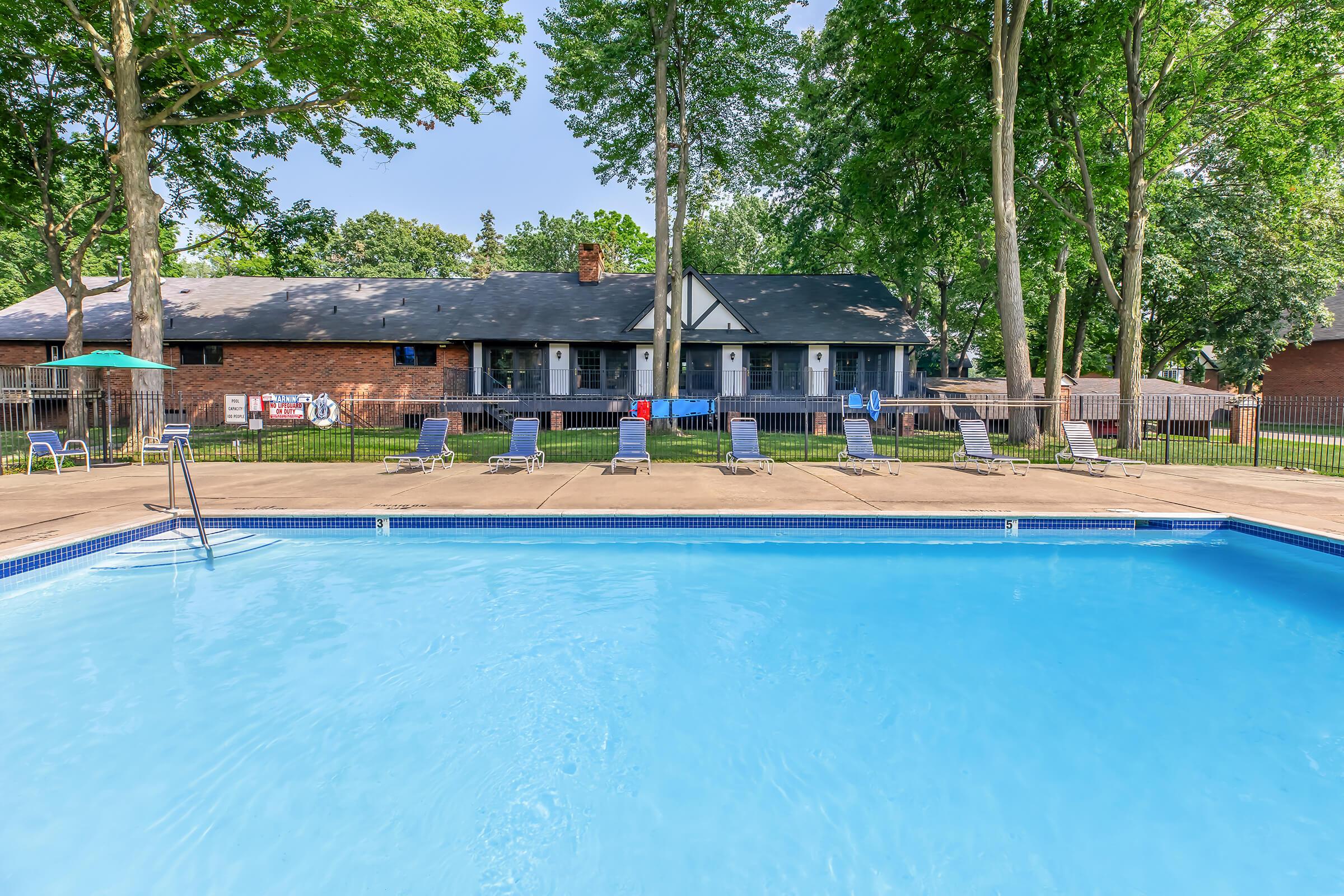 A serene swimming pool surrounded by lounge chairs, with a lush green lawn and tall trees. In the background, a brick building with multiple doors is visible, and there are umbrellas by the poolside. The scene is bright and inviting, perfect for relaxation and leisure activities.
