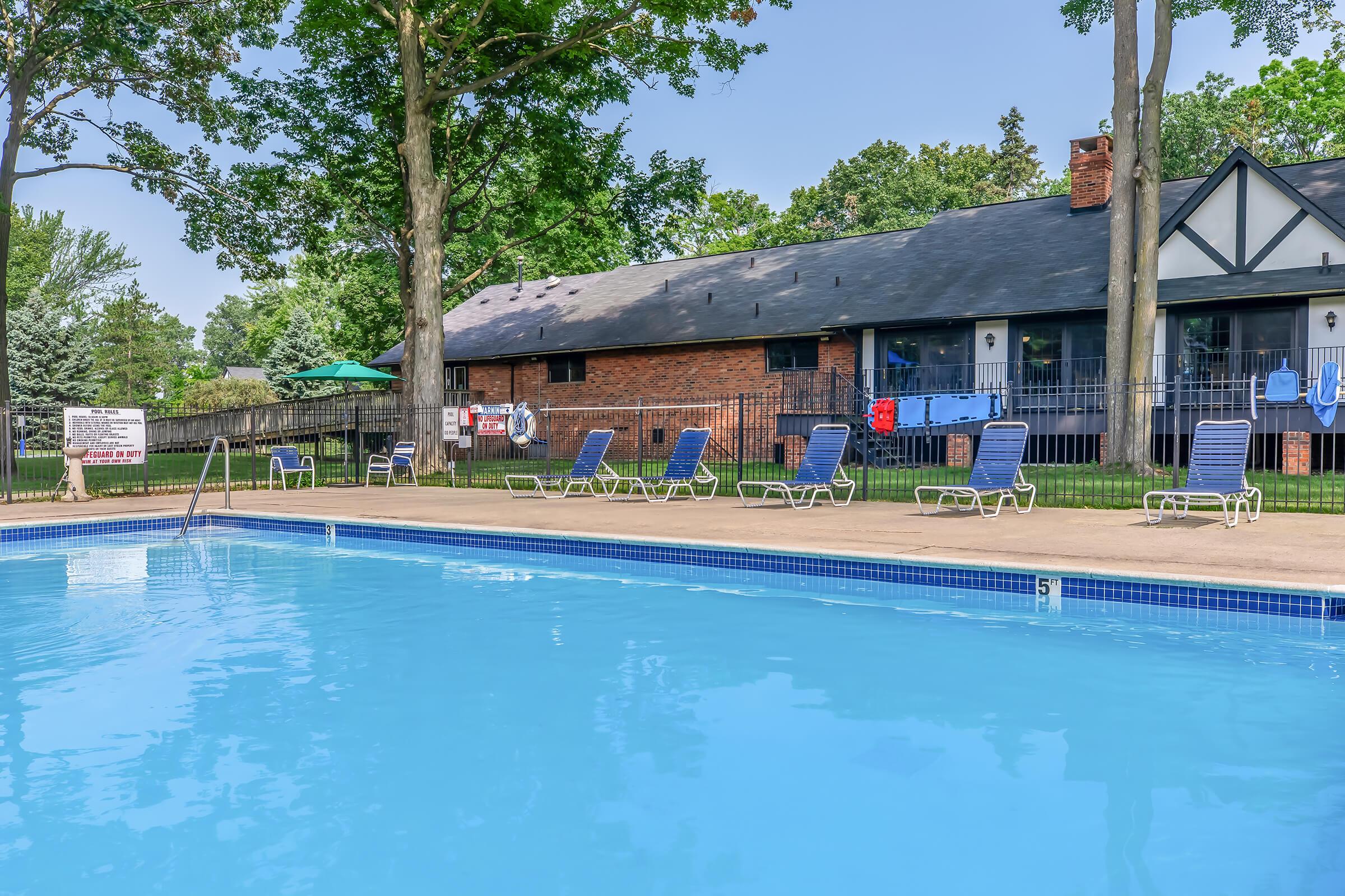 A clear blue swimming pool surrounded by lounge chairs, with trees in the background. A brick building with a black roof is seen to the right, and a green umbrella is visible nearby. Poolside signs indicate safety information. The scene is bright and inviting, perfect for relaxation.