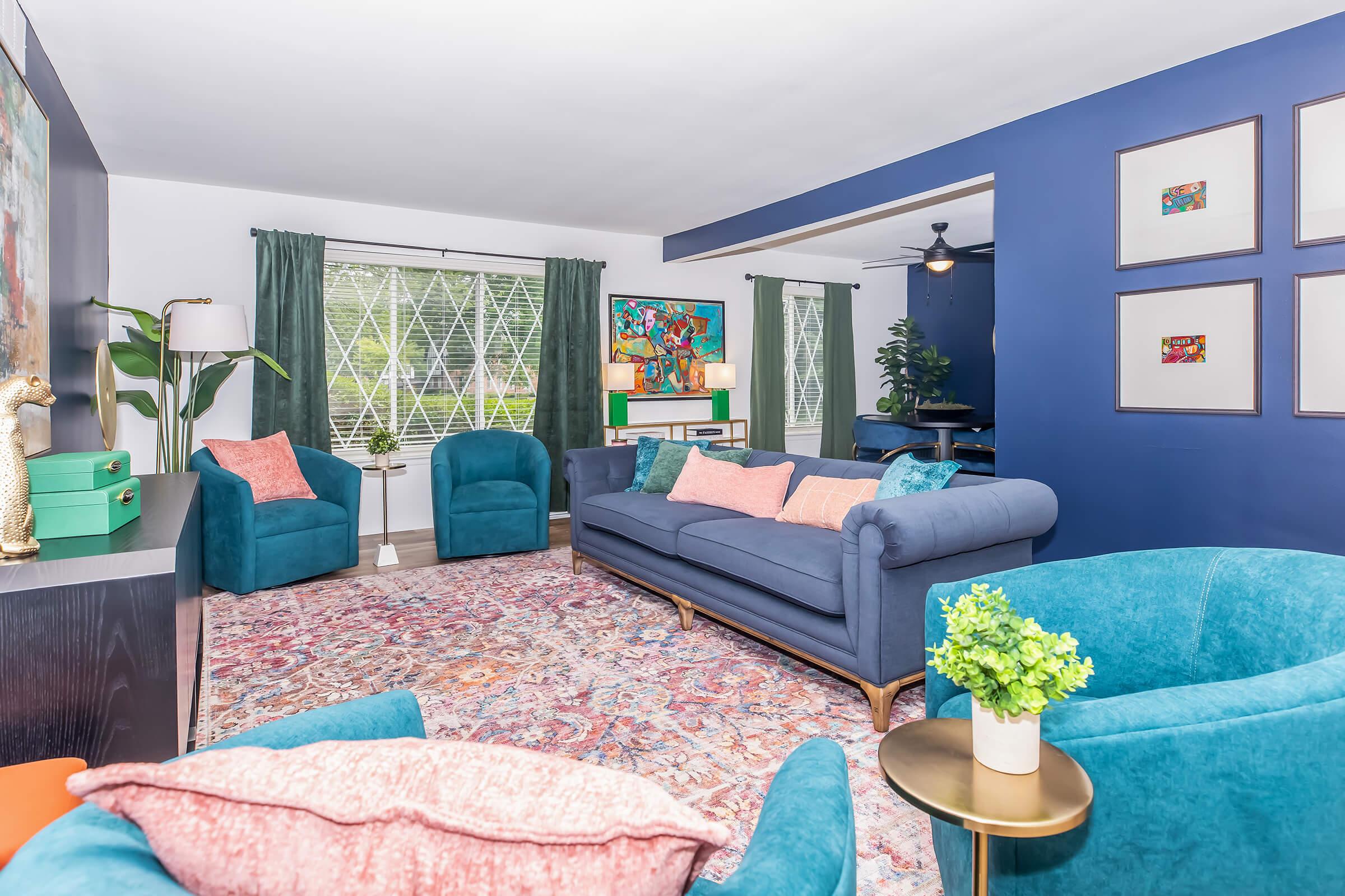 A stylish living room featuring a blue sofa with colorful pillows, two teal armchairs, and a vibrant area rug. Large windows with green drapes allow natural light, complemented by a green plant and artwork on the walls. A stylish side table adds elegance to the cozy space.