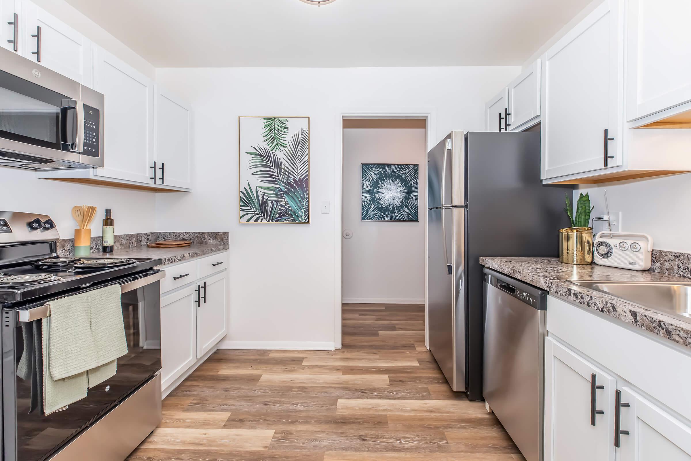 A modern kitchen featuring white cabinetry, stainless steel appliances, and granite countertops. The space includes a stove, refrigerator, and dishwasher, with wooden flooring. Two wall art pieces depict plant themes, adding a decorative touch to the bright, inviting atmosphere.