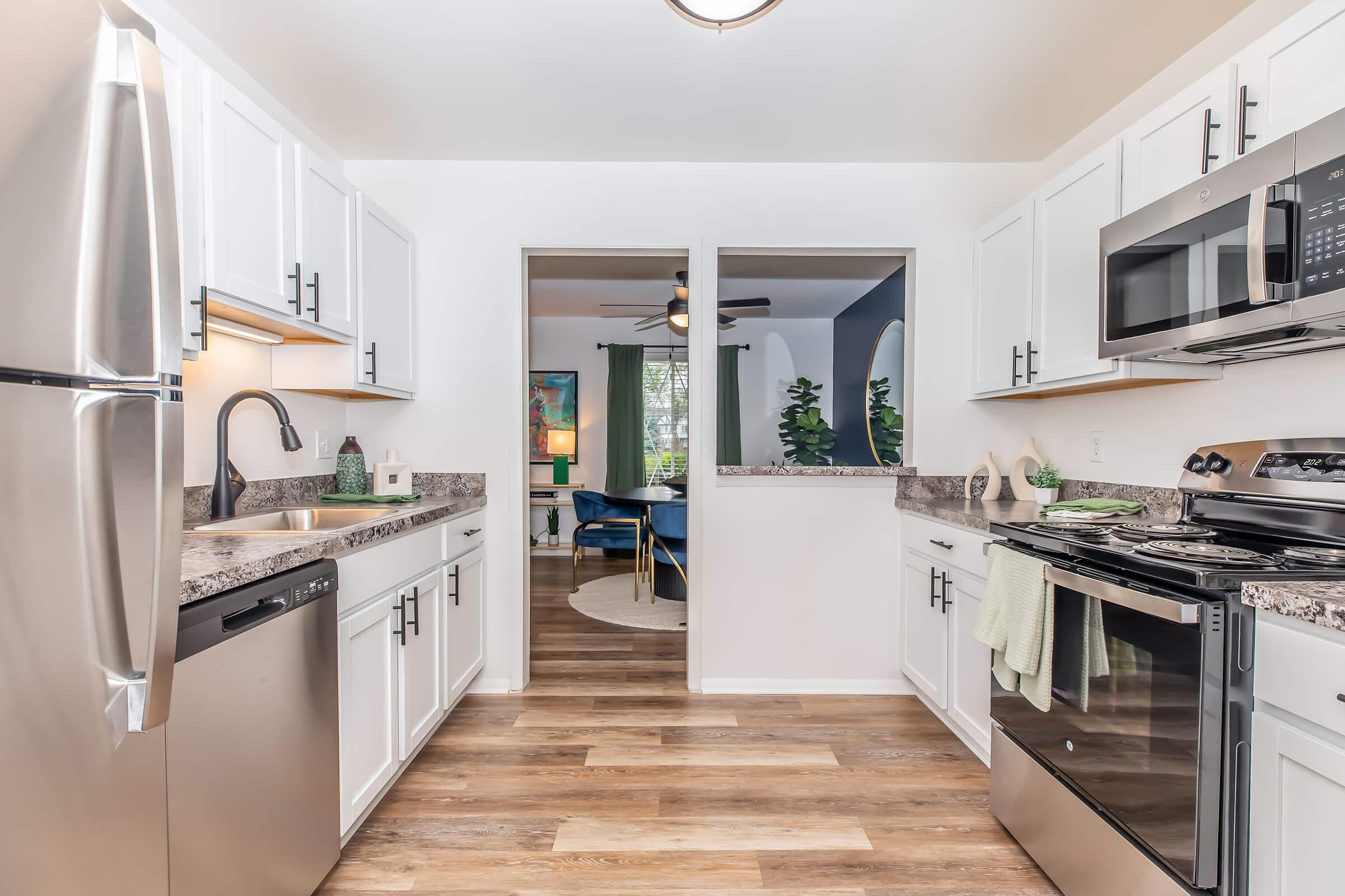 A modern kitchen featuring white cabinets, stainless steel appliances, and granite countertops. The space includes a dishwasher, stove, and microwave, with a view leading into a living area. Natural light illuminates the room, highlighting the wood-like flooring. Green accents and decor enhance the inviting atmosphere.