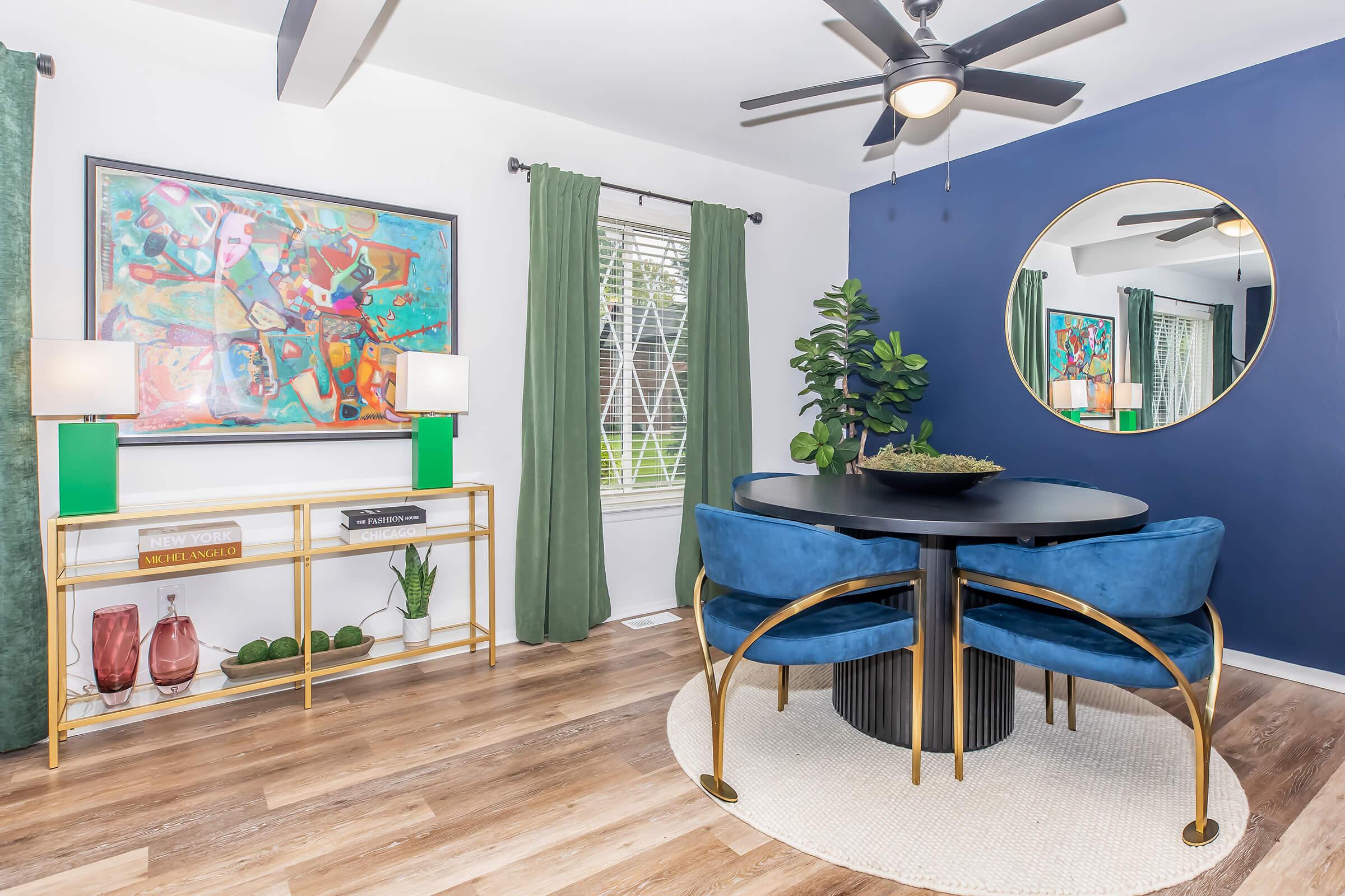 A cozy dining area featuring a round black table with blue chairs, a potted plant, and decorative items. The walls are painted in contrasting shades of blue and white, with green curtains framing a window. An abstract painting adorns the wall above the table, complemented by stylish lamps on a gold shelf.