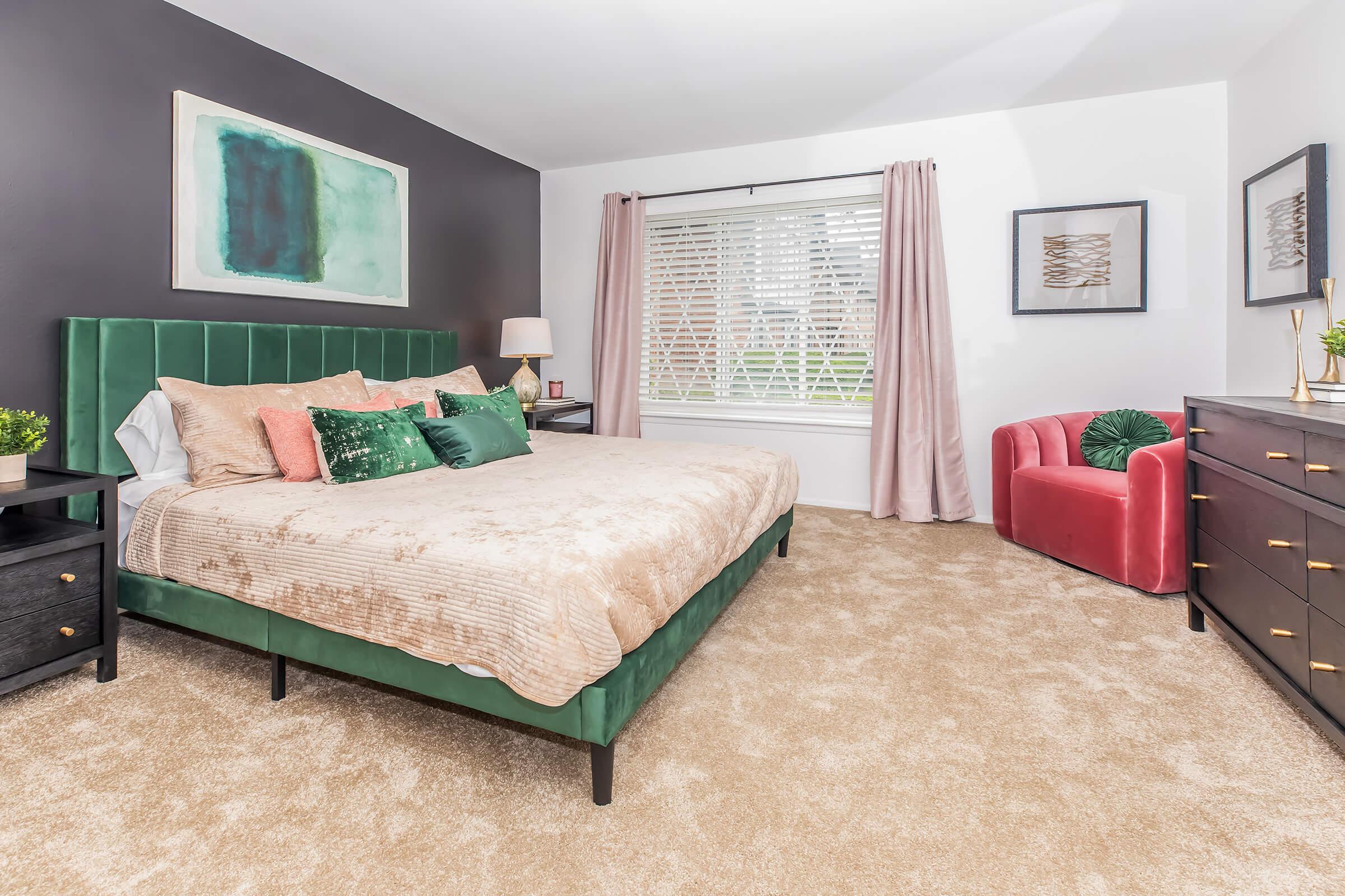 A modern bedroom featuring a green upholstered bed with decorative pillows, a nightstand with a lamp, and a plush carpet. A window with white blinds is adorned with sheer curtains. To the right, there's a cozy red chair and a dark wood dresser. Wall art and decorative plants enhance the stylish decor.