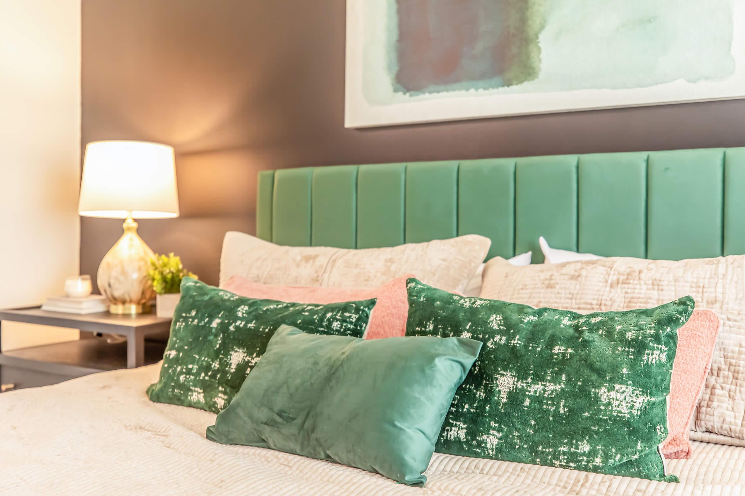 A cozy bedroom scene featuring a green and pink color scheme. The bed is adorned with plush pillows in various shades of green and pink. A stylish lamp casts a warm glow, and artwork in calming colors is mounted on the wall above the headboard. A small plant and bedside table complete the inviting atmosphere.