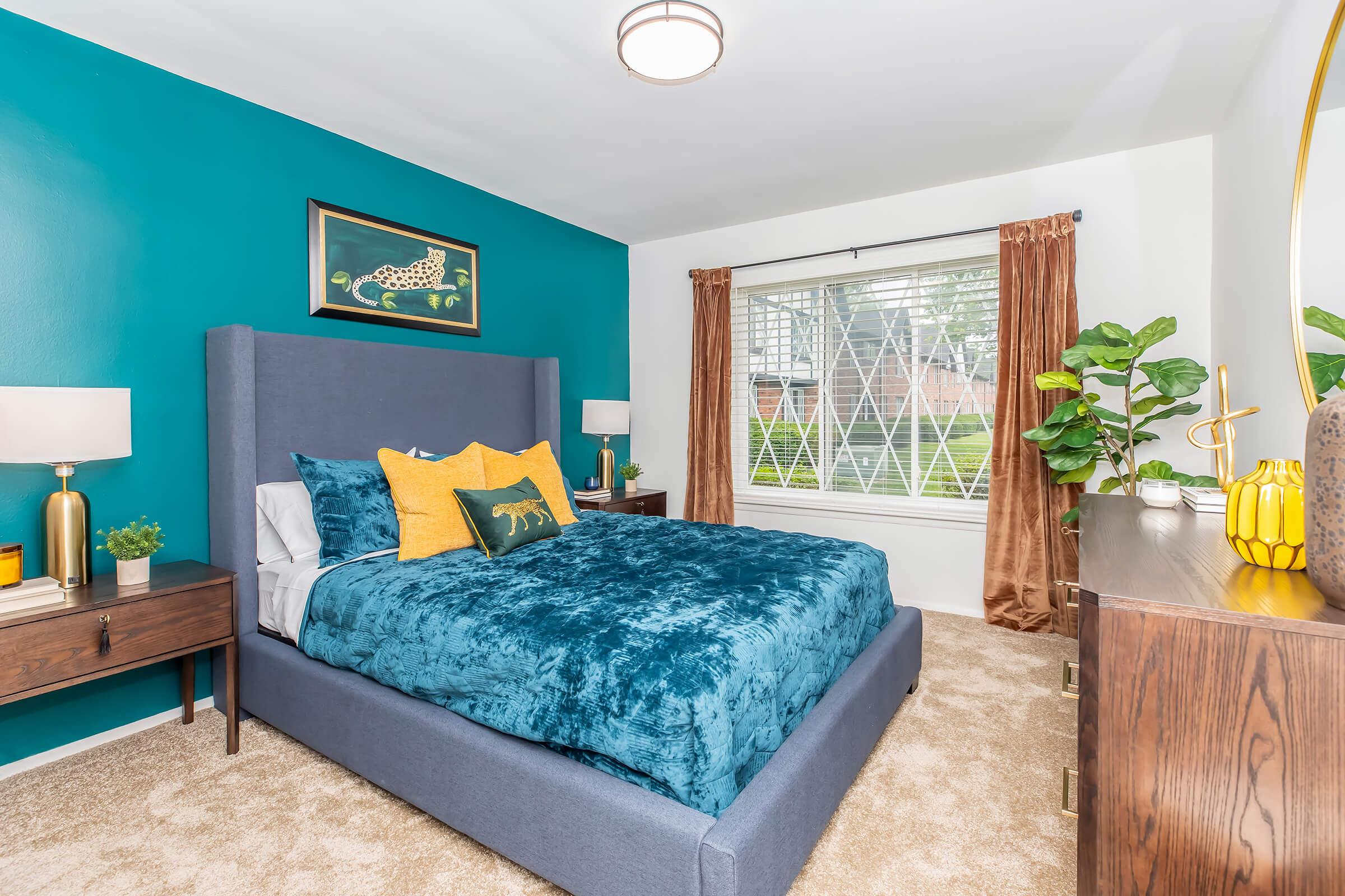 A cozy bedroom featuring a blue upholstered bed with vibrant yellow and teal pillows. The walls are painted a deep teal, with a large window adorned by brown curtains, allowing natural light in. A bedside table holds decorative lamps and plants, while a stylish dresser completes the inviting space.