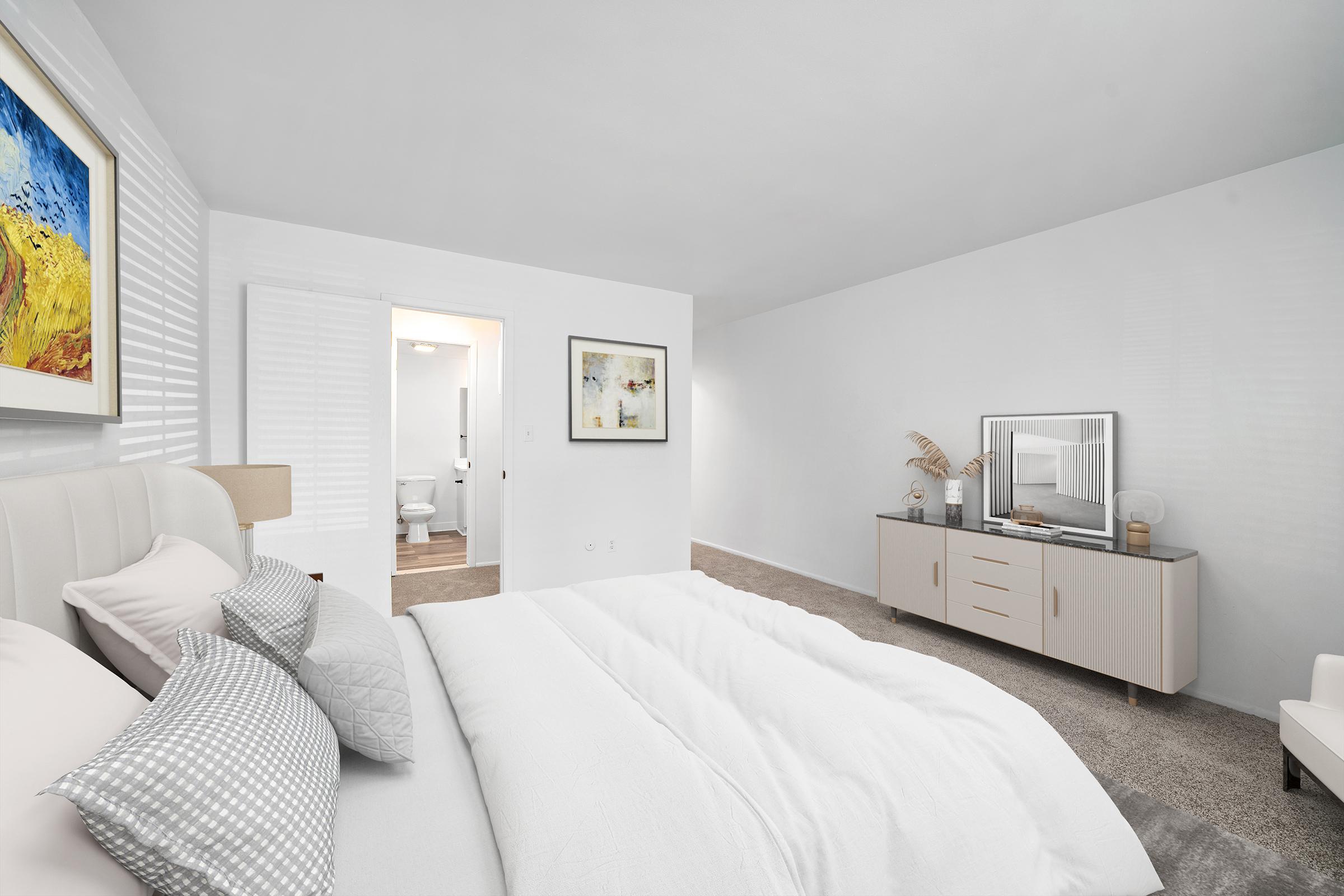 A modern bedroom featuring a neatly made bed with white linens and decorative pillows. There's a light-colored dresser with a mirror, a chair in the corner, and artwork on the wall. A door leading to a bathroom is visible in the background, and the room has light-colored walls and carpet.