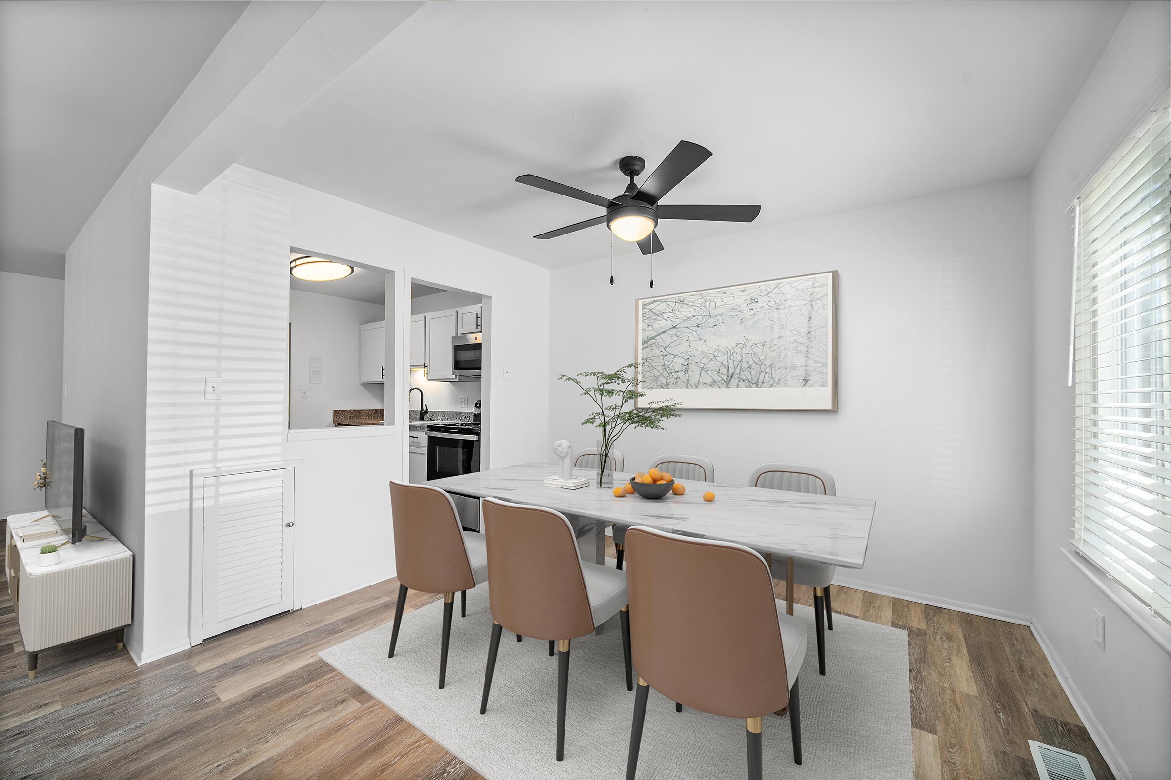 A modern dining area featuring a large marble table surrounded by brown chairs. A ceiling fan hangs above, and a painting of trees is displayed on the white wall. A small plant and a bowl of oranges sit on the table. The room has a light, airy feel with large windows allowing natural light to enter.