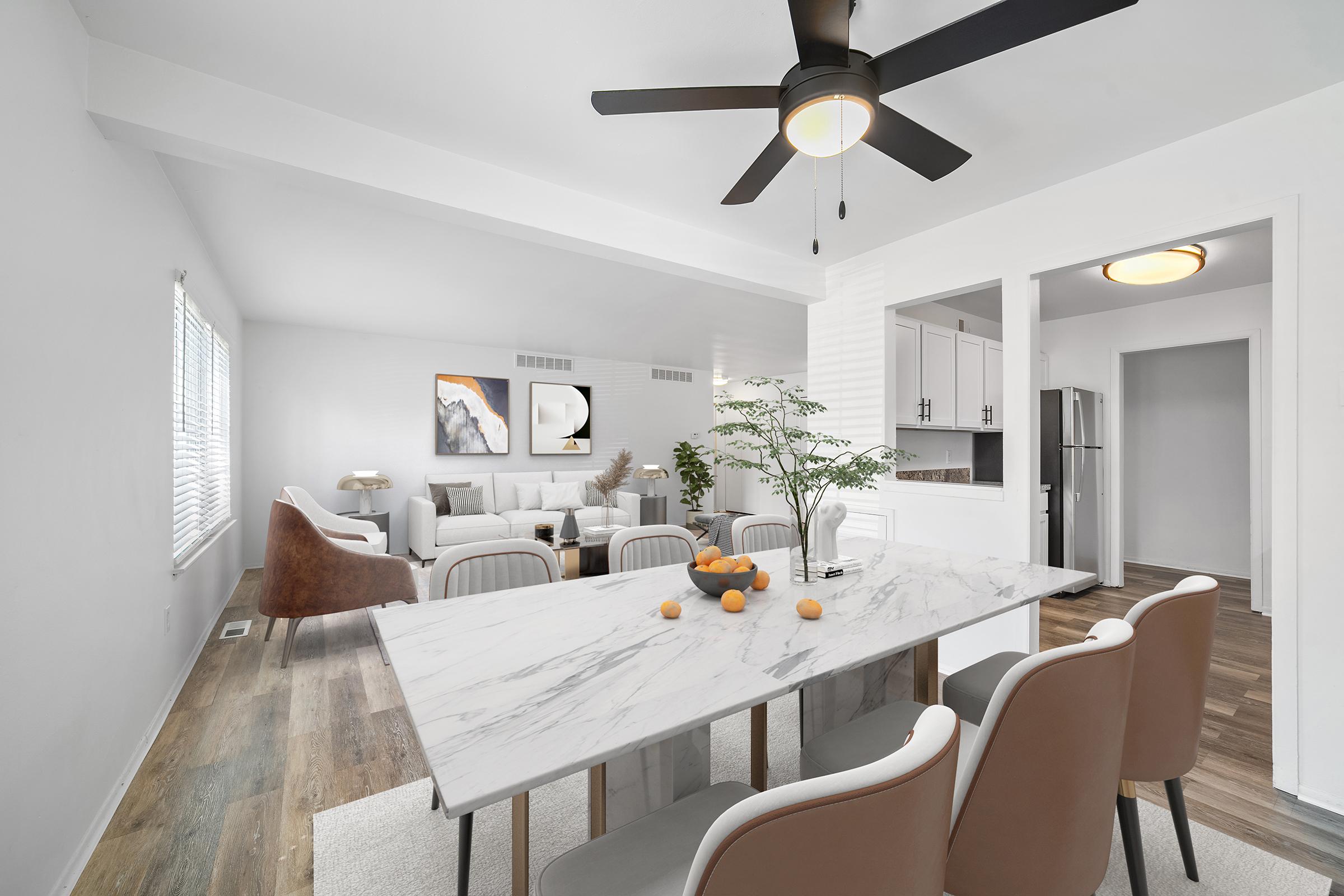 A modern, bright living and dining area featuring a marble dining table with brown chairs, a white sofa, and neutral decor. The space is adorned with plants and artwork on the walls, complemented by a ceiling fan. A kitchen area is visible in the background, creating an inviting atmosphere.