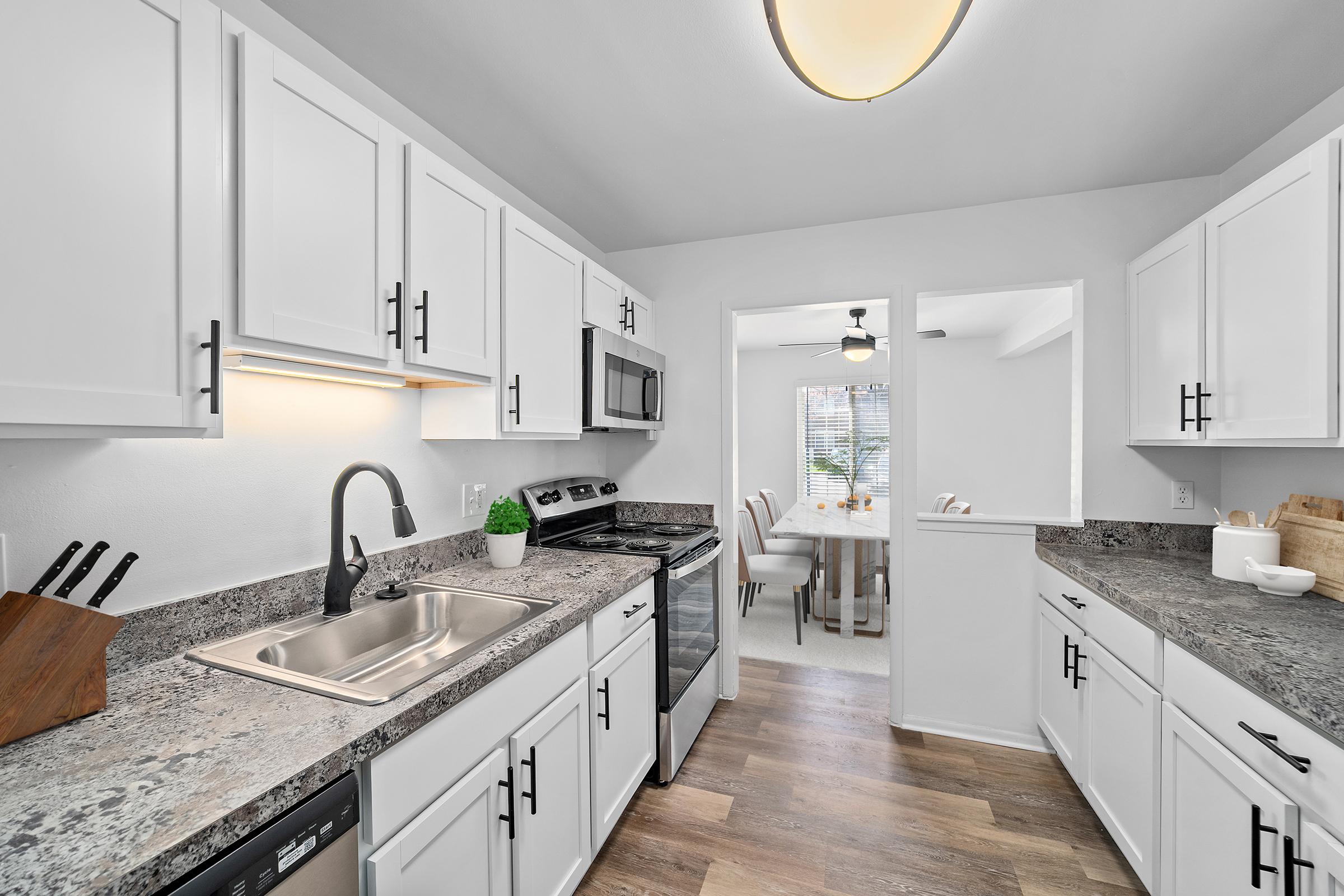 A modern kitchen featuring white cabinets, stainless steel appliances, and a granite countertop. The sink is placed beneath a window with natural light. A dining area with a ceiling fan is visible in the background, creating an open and inviting space.