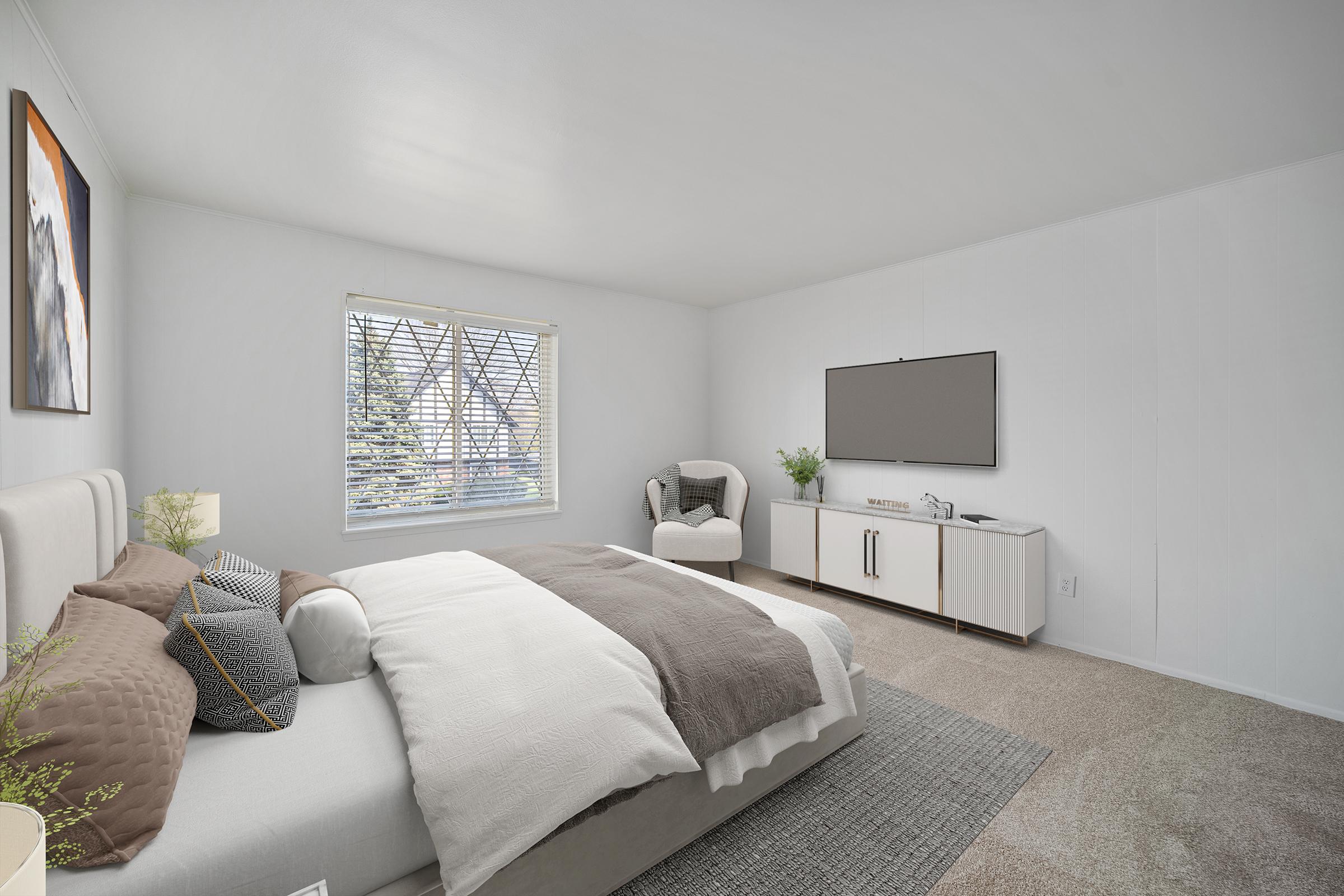 A spacious, modern bedroom featuring a large bed with neutral-toned bedding, a cozy armchair, and a flat-screen TV mounted on the wall. Natural light streams in through a window adorned with blinds, and a small console table holds decorative items and a plant, creating a serene and inviting atmosphere.