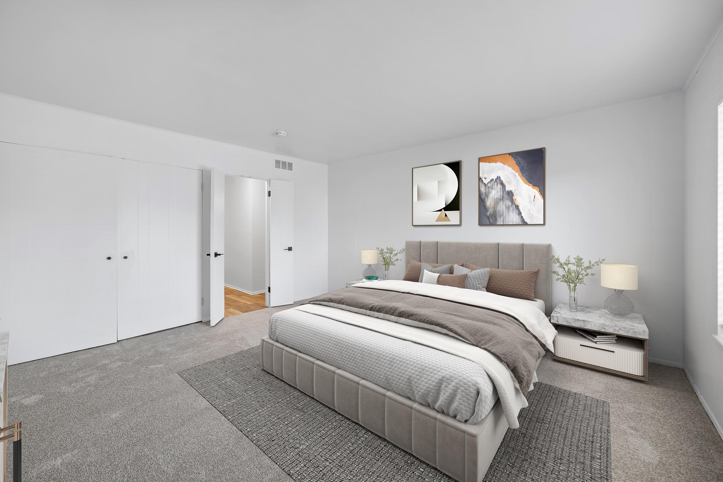 A modern bedroom featuring a large bed with a soft gray headboard and layered bedding. Two framed artworks hang on the wall above the bed. A bedside table with a lamp and small plants is on one side. A large area rug covers the floor, and there are white closet doors and a doorway leading to another space.