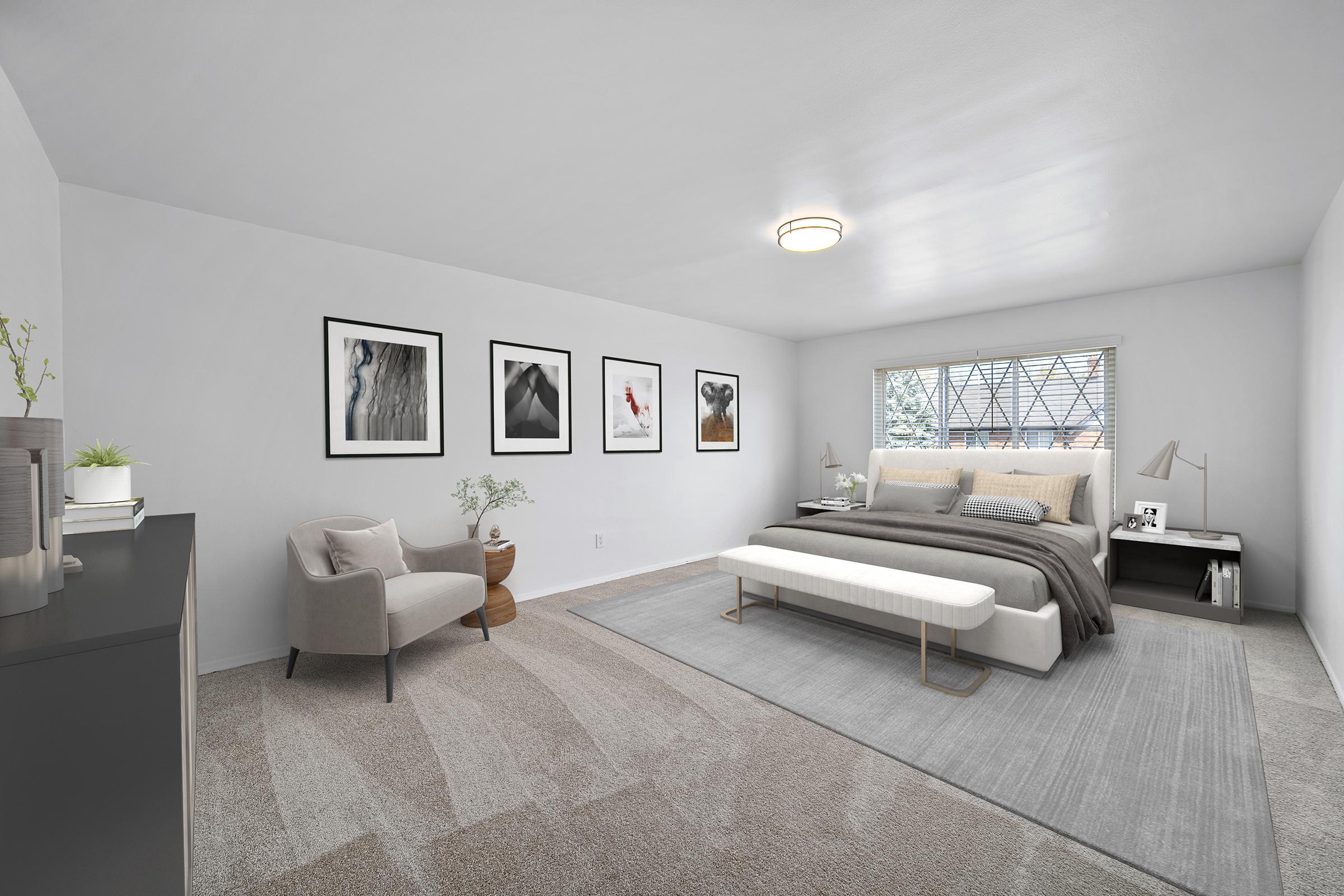 A modern bedroom featuring a king-size bed with neutral bedding and a bench at the foot. Two artful prints hang on the wall above the bed. A cozy armchair sits in one corner, with a small plant and decorative items on a dark dresser. A large window lets in natural light, illuminating the soft carpet and warm color palette.