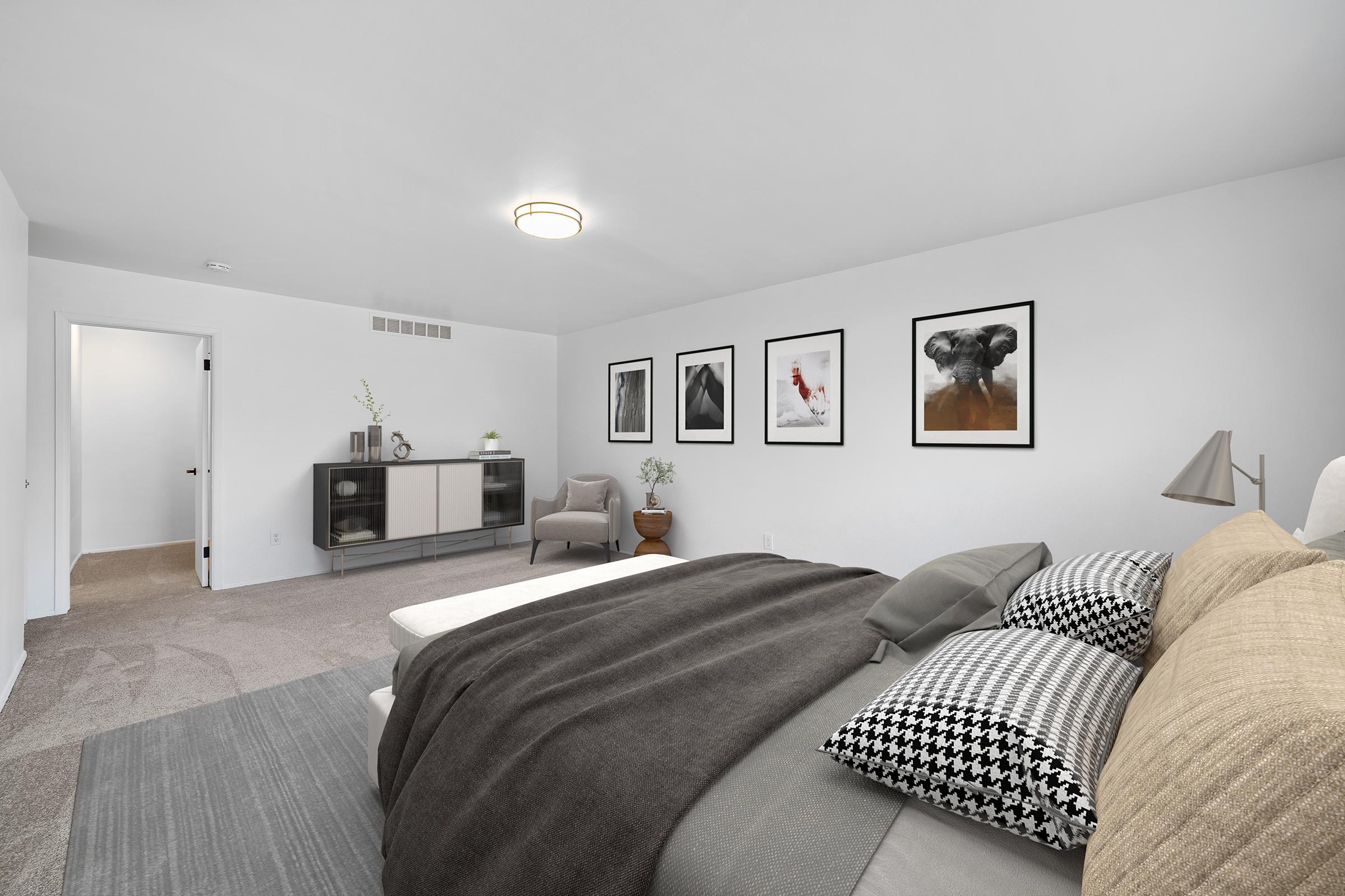A modern bedroom featuring a neatly made bed with gray and patterned pillows, a cozy throw blanket, and a gray area rug. The walls are adorned with framed art prints, and a stylish cabinet is placed opposite the bed. A comfortable armchair and a potted plant complete the inviting atmosphere.