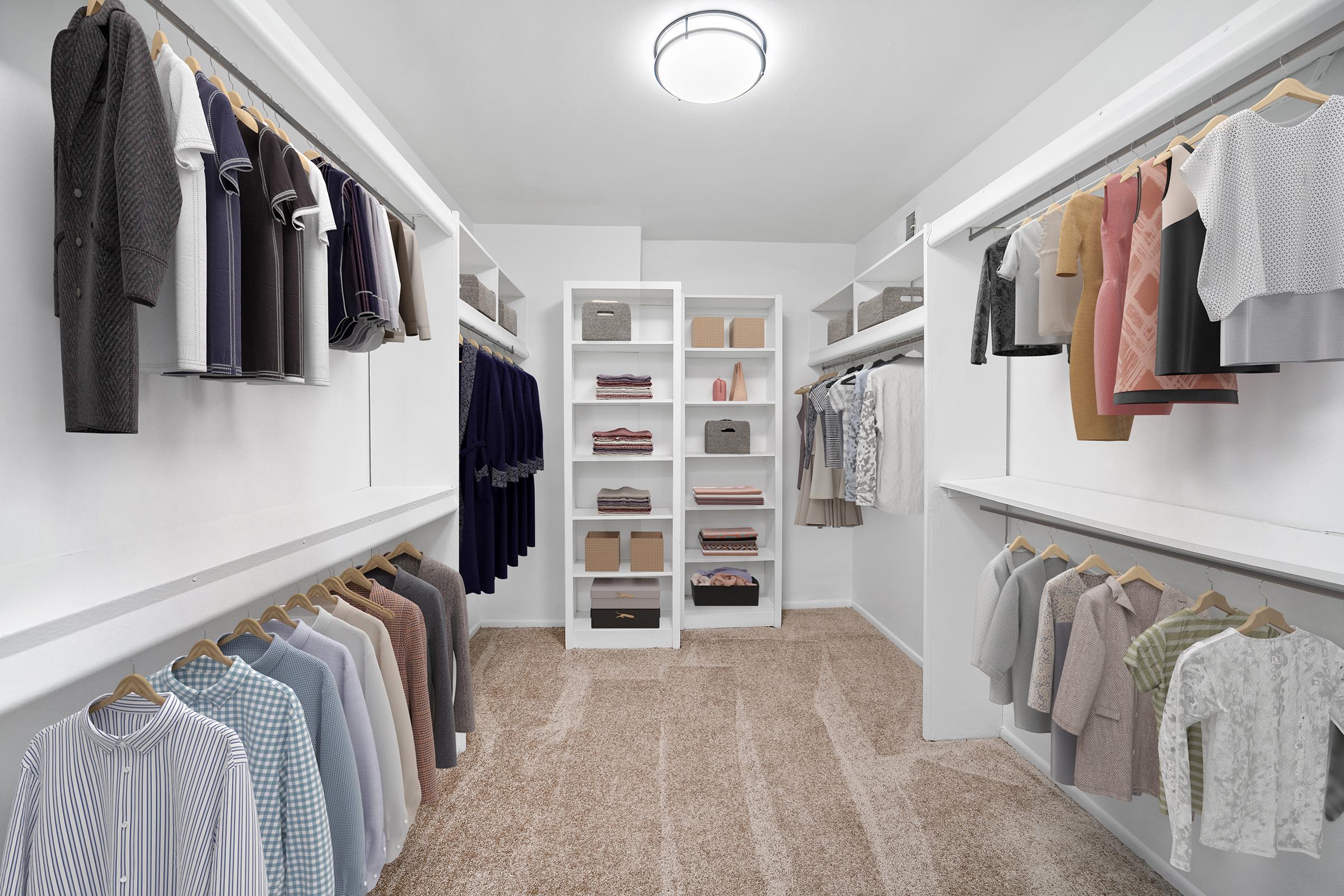A neatly organized walk-in closet featuring hanging clothes on both sides, including shirts, blouses, and coats. On the back wall, there are shelves storing items in boxes and folded garments. The flooring is carpeted, and overhead lighting provides bright illumination. The overall aesthetic is clean and modern.