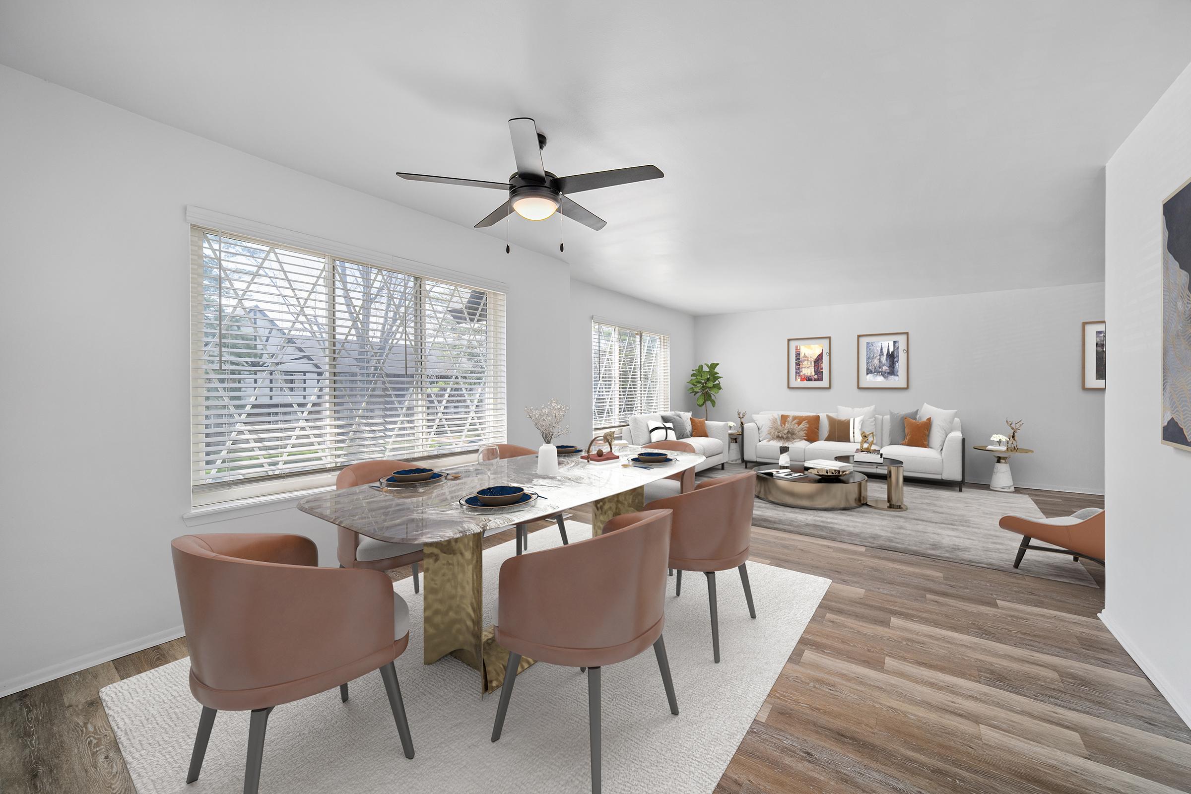 A modern dining area featuring a large marble table with brown chairs, situated next to a bright living space. The room has a ceiling fan and large windows letting in natural light. The decor is minimalistic, with art pieces on the walls and a light-colored rug under the dining table, creating an inviting atmosphere.
