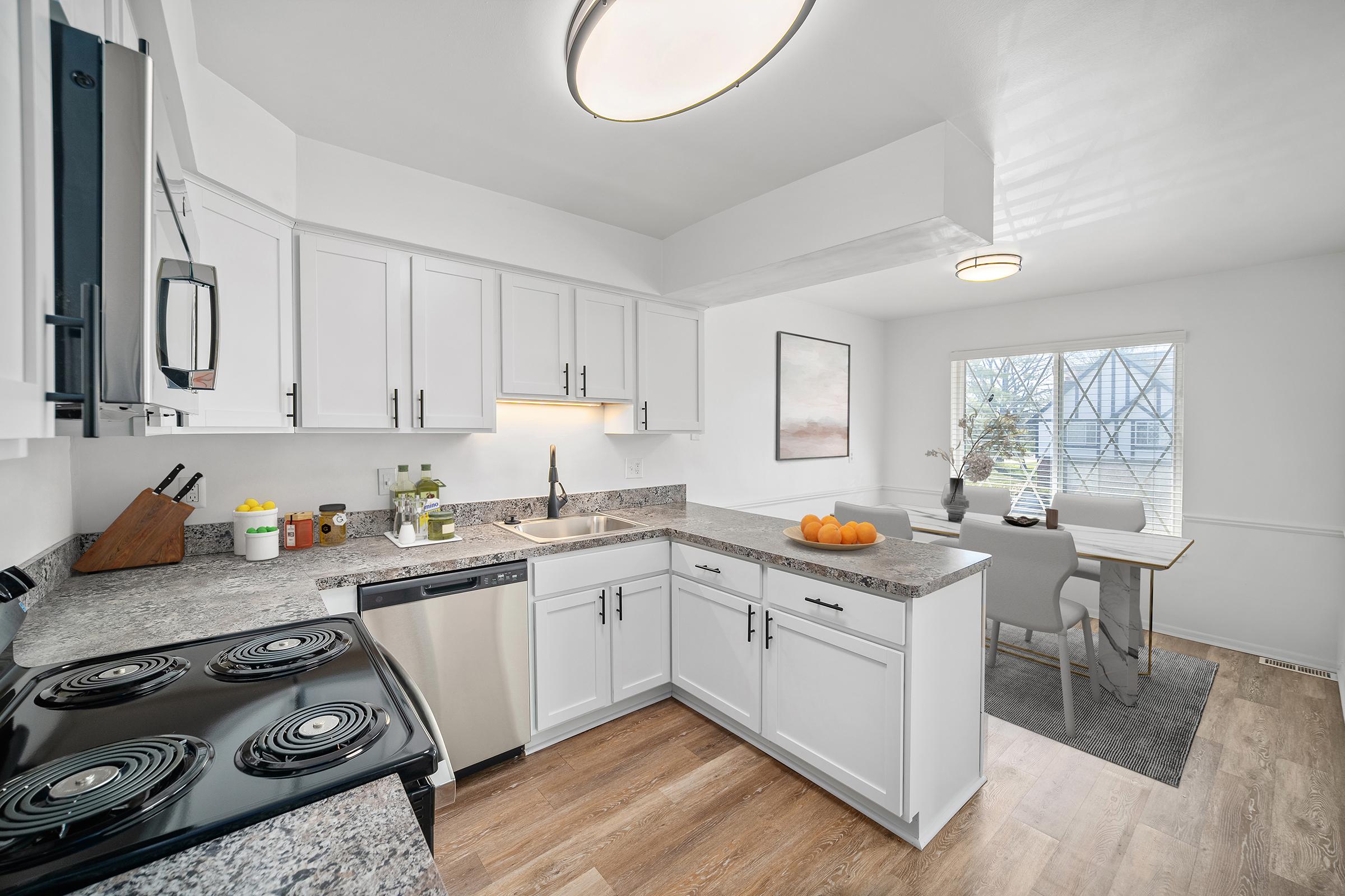 Bright and modern kitchen featuring white cabinets, a granite countertop, and stainless steel appliances. A black stove is visible, along with a sink and decorative items like fruits. A dining area with a table and chairs is situated near a window, allowing natural light to fill the space.