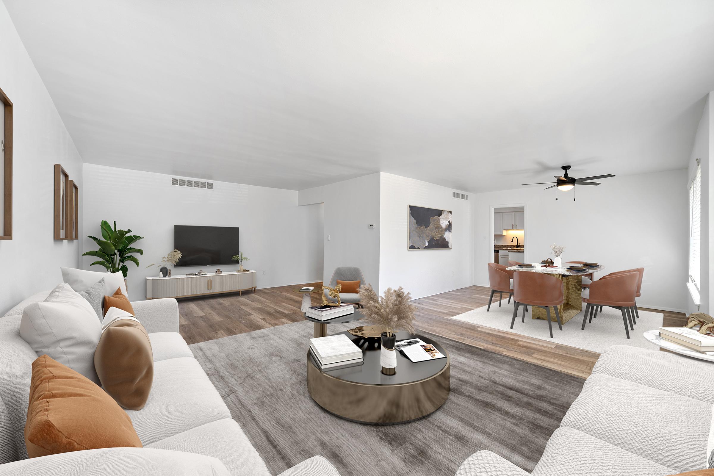 A modern living room featuring a light color scheme with a white sofa and accent pillows in shades of orange. There is a round coffee table with decorative items, a flat-screen TV mounted on the wall, and a cozy seating area with tan chairs. The space is bright and airy, with a ceiling fan and wooden flooring.