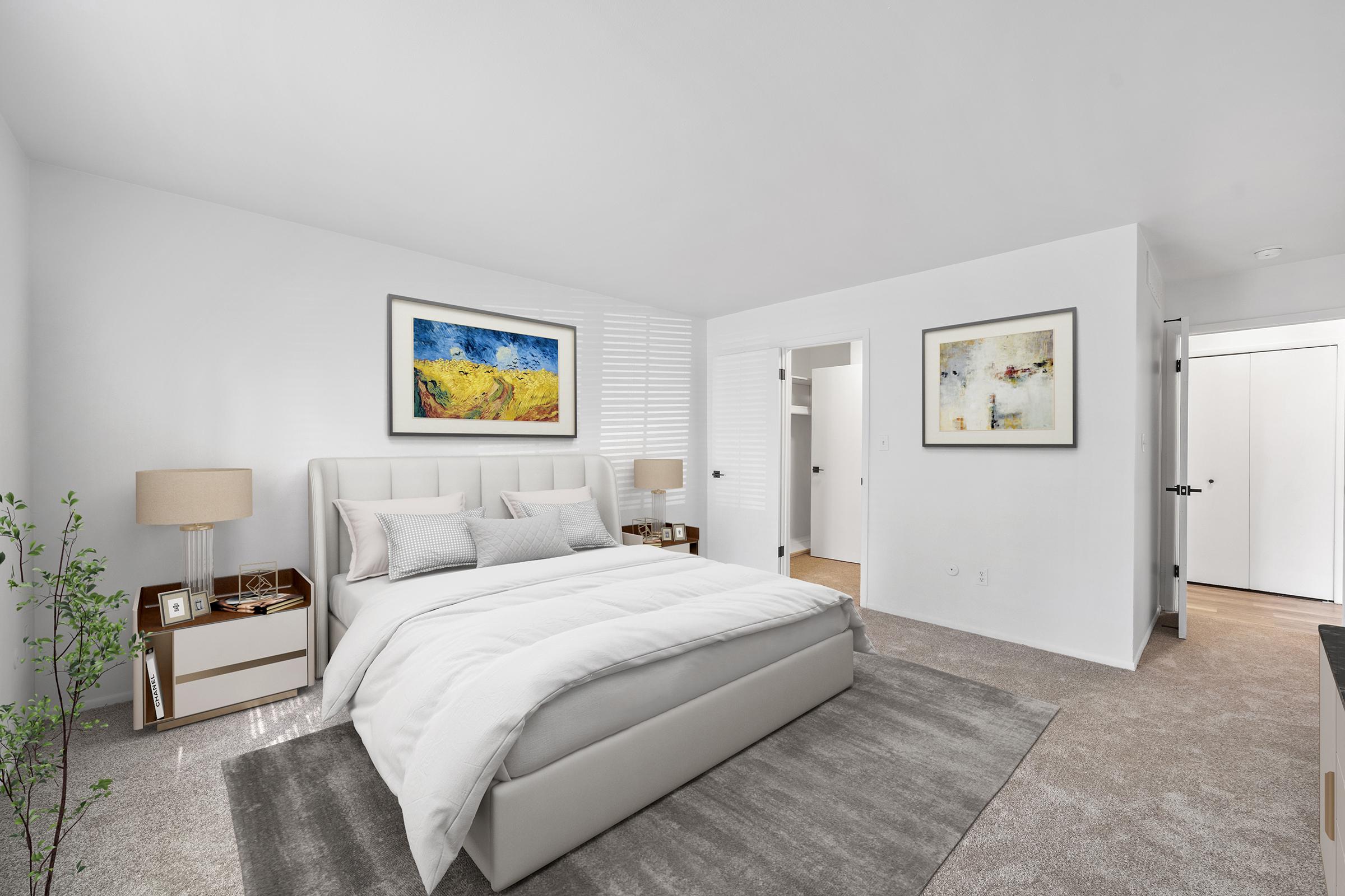 A bright and modern bedroom featuring a large bed with a light gray upholstered headboard, white bedding, and decorative pillows. Two bedside tables with lamps are on either side. Decorative artwork hangs on the white walls, and there is a plush area rug on the carpeted floor. A door leads to a closet and another room.