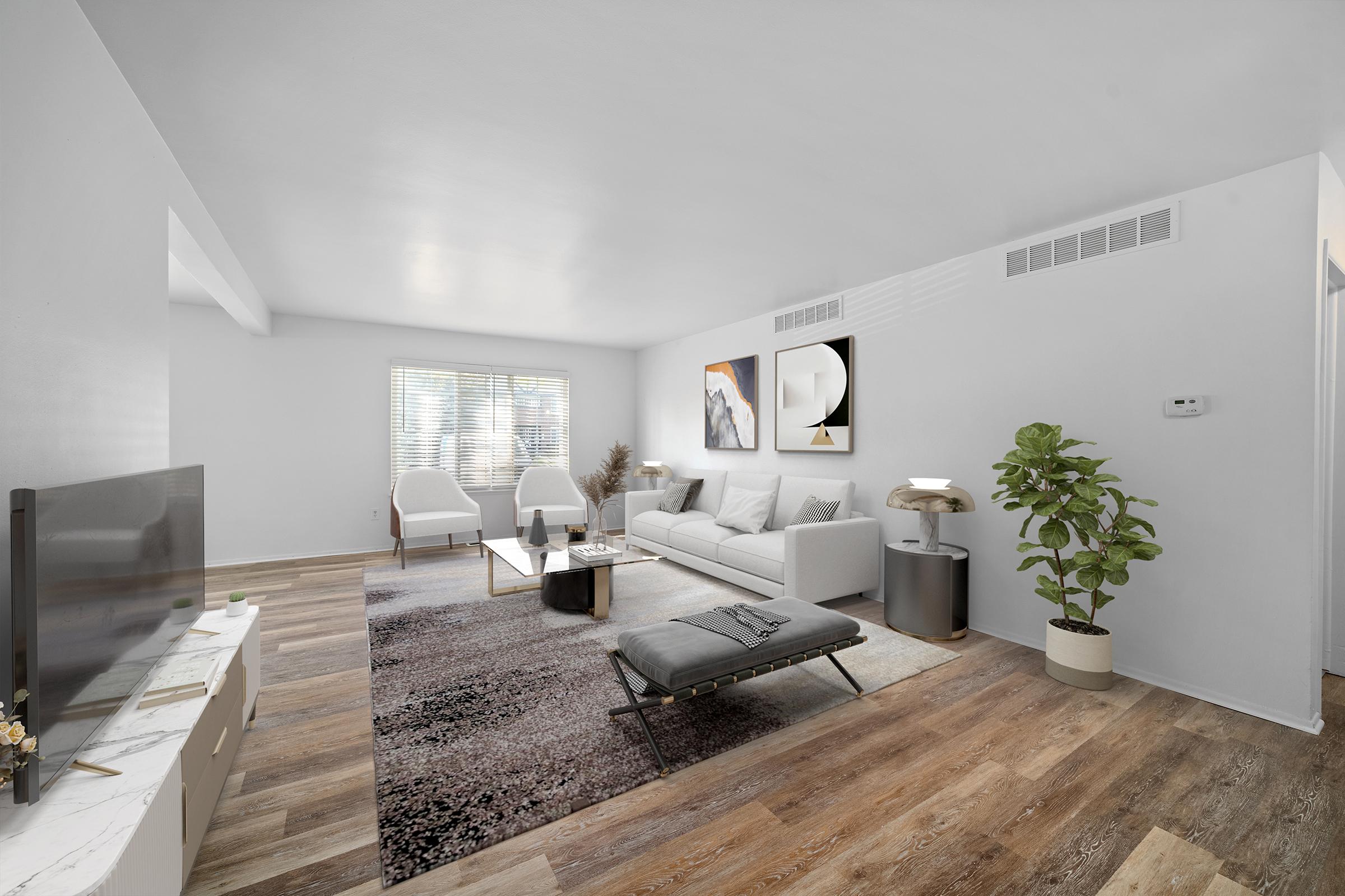 A modern, stylish living room featuring a light gray sofa, two white armchairs, and a coffee table on a patterned rug. Natural light streams through a window, illuminating a minimalist decor with a leafy plant and decorative wall art. The flooring is a warm wood finish, creating a cozy atmosphere.