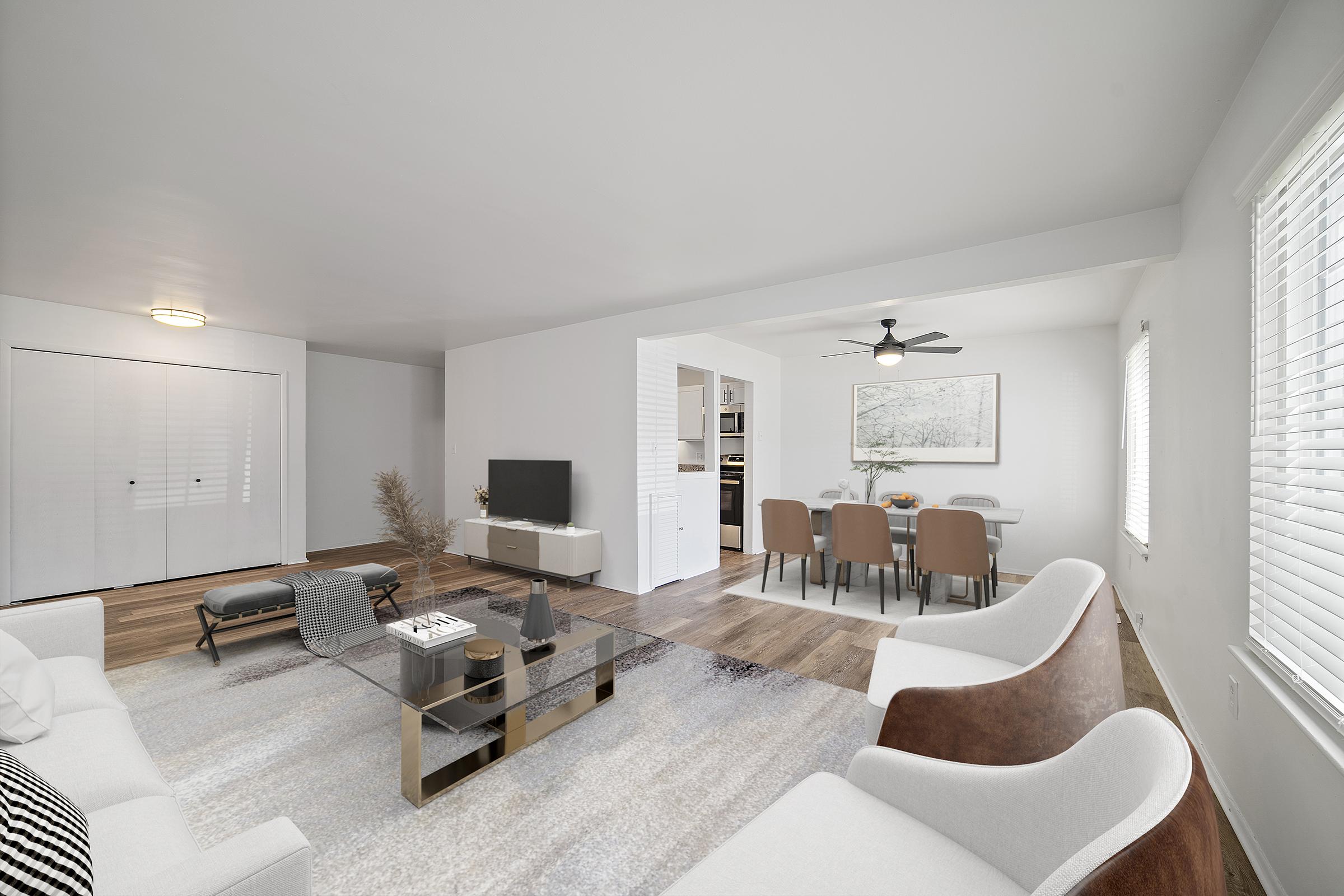 A modern, well-lit living room featuring a minimalist design. It includes a white sofa, stylish armchairs, a glass coffee table on a soft rug, and a flat-screen TV. A dining area with a table and chairs is visible in the background, along with a ceiling fan and large windows allowing natural light.