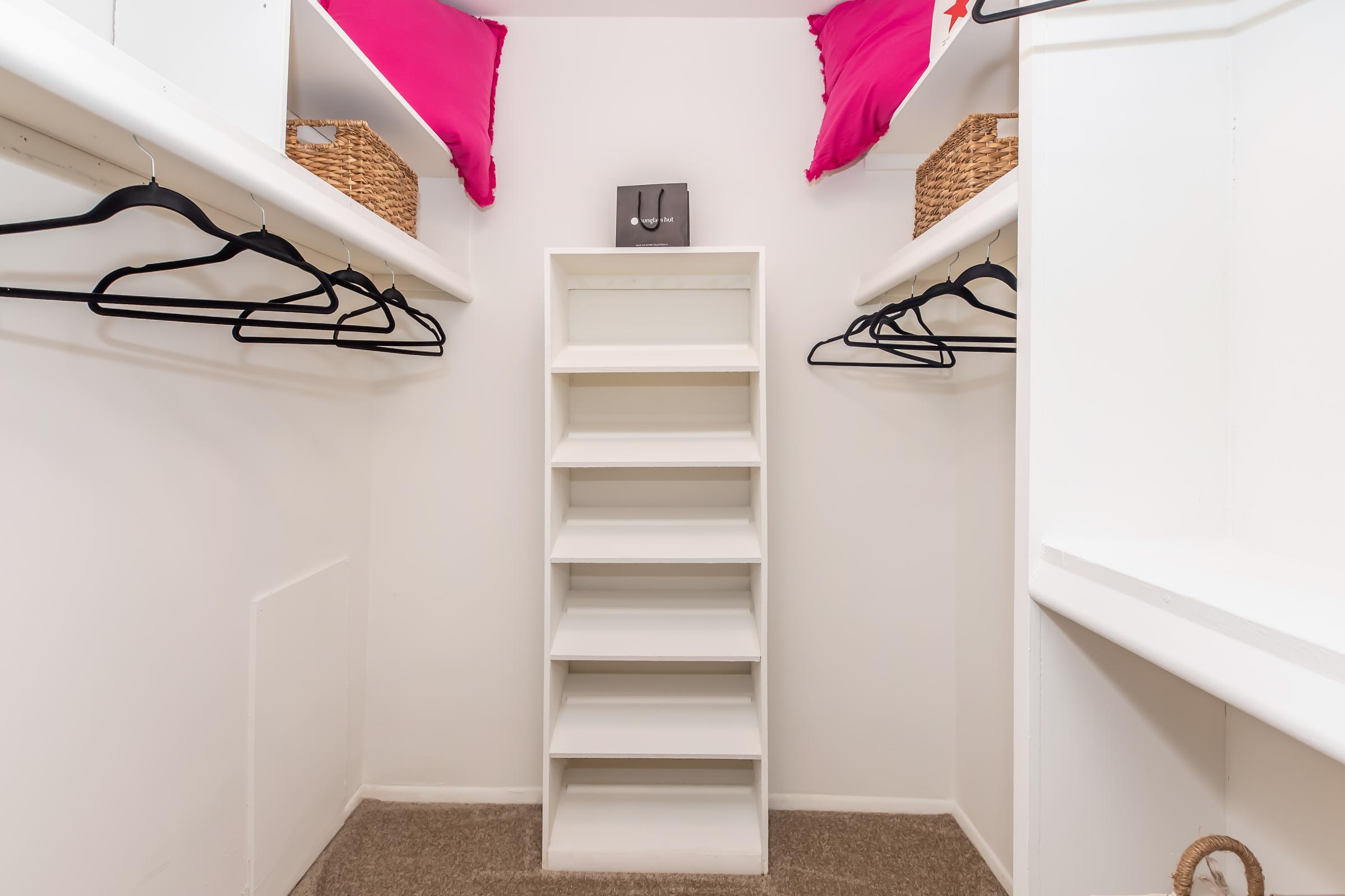 A neatly organized closet featuring white shelving units and several empty hangers. Two woven baskets are placed on top shelves, and pink pillows are positioned on the upper shelf, creating a tidy and functional storage space. The floor is carpeted, adding a cozy touch.