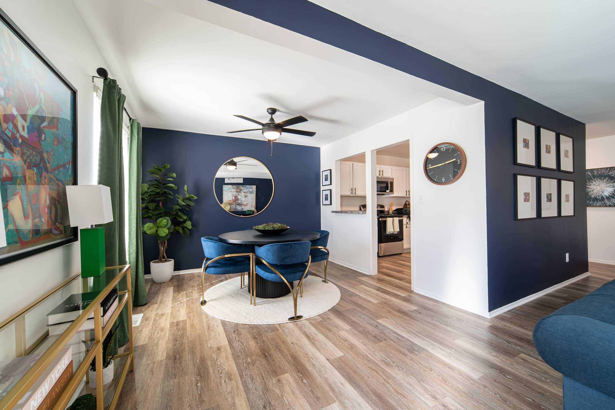 A modern dining area with a round table surrounded by blue velvet chairs. A circular mirror hangs on the dark blue wall, reflecting light. There's a green plant in the corner, and a light fixture on the ceiling. The room features wooden flooring and is open to a kitchen area in the background.