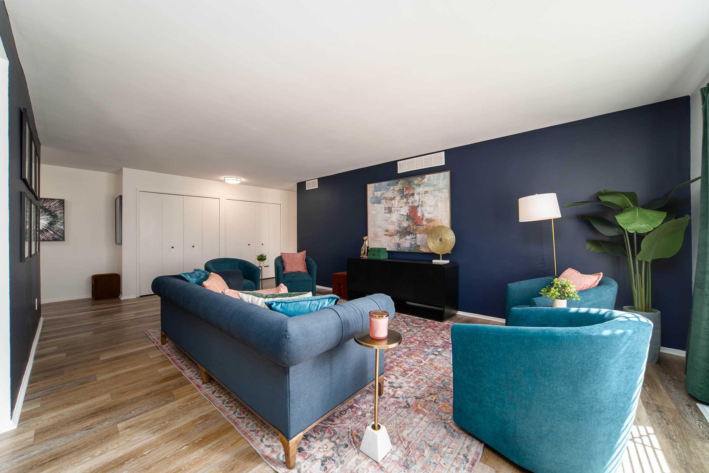 A modern living room featuring plush teal sofas, decorative pillows, and a vibrant area rug. The walls are painted dark blue, complemented by a colorful abstract artwork. There's a stylish lamp, a plant in the corner, and a sleek black console table, creating a cozy and inviting atmosphere.