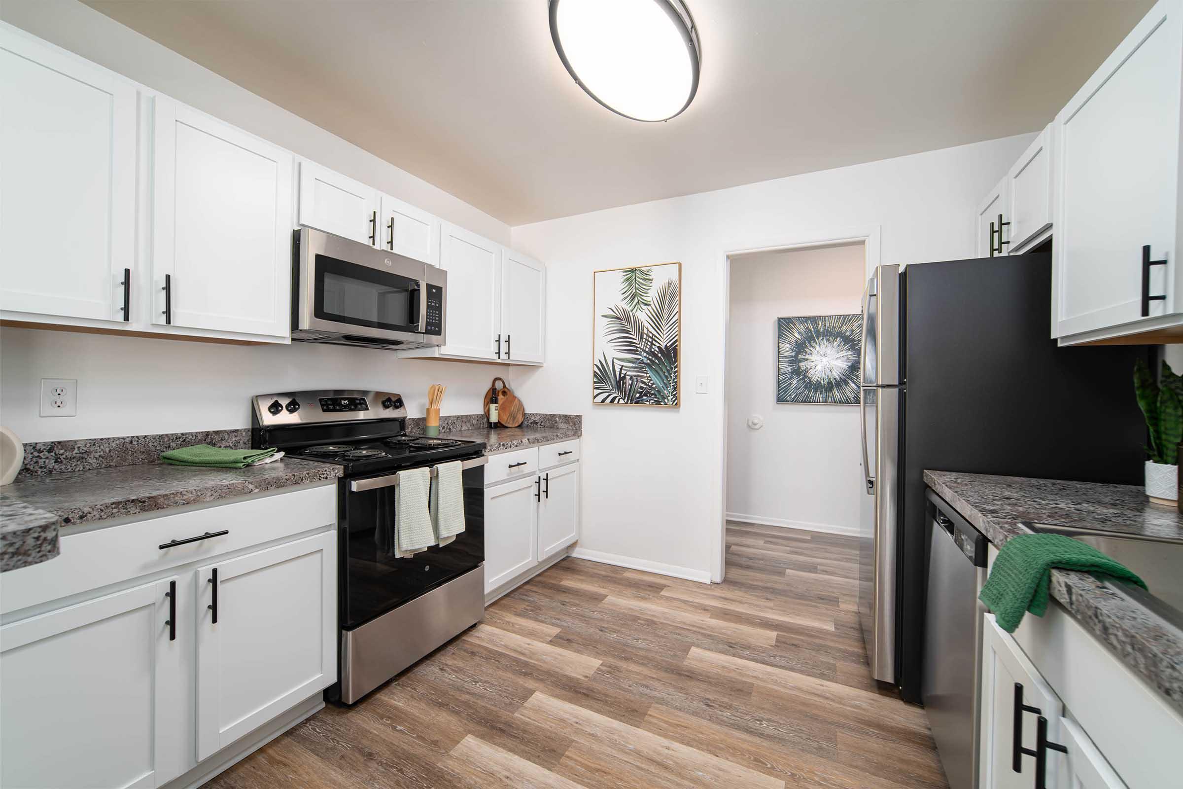 A modern kitchen featuring white cabinets, stainless steel appliances, and a granite countertop. The space includes a microwave, oven, and refrigerator, with green accents in the form of towels and decor. Two framed artworks with nature themes adorn the walls, enhancing the inviting atmosphere.