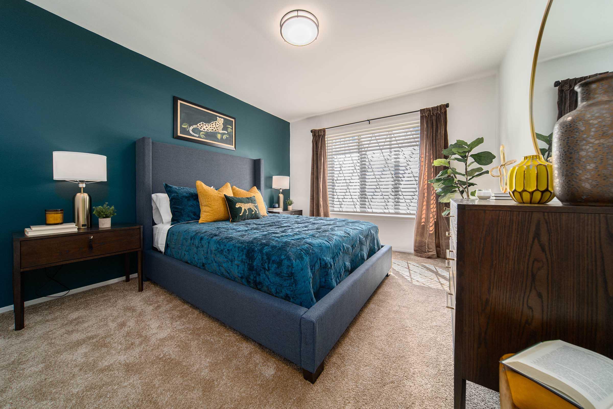 A cozy bedroom featuring a blue accent wall, a plush bed with teal and yellow bedding, two bedside lamps, and a wooden nightstand. Natural light filters through a window adorned with dark curtains. A decorative plant and a stylish vase add a touch of greenery and elegance to the space.