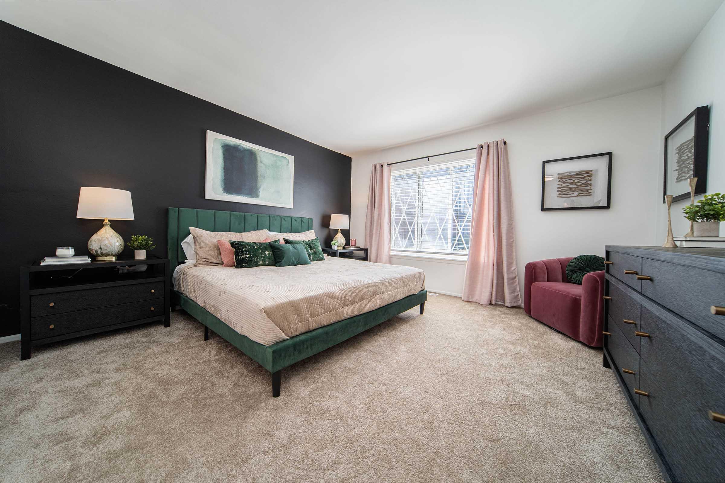 A cozy bedroom featuring a green upholstered bed with decorative pillows, a black accent wall, and a large window with pink curtains. There’s a bedside table with a lamp, a dark wood dresser, and a burgundy armchair in the corner. Soft carpeting completes the inviting atmosphere.