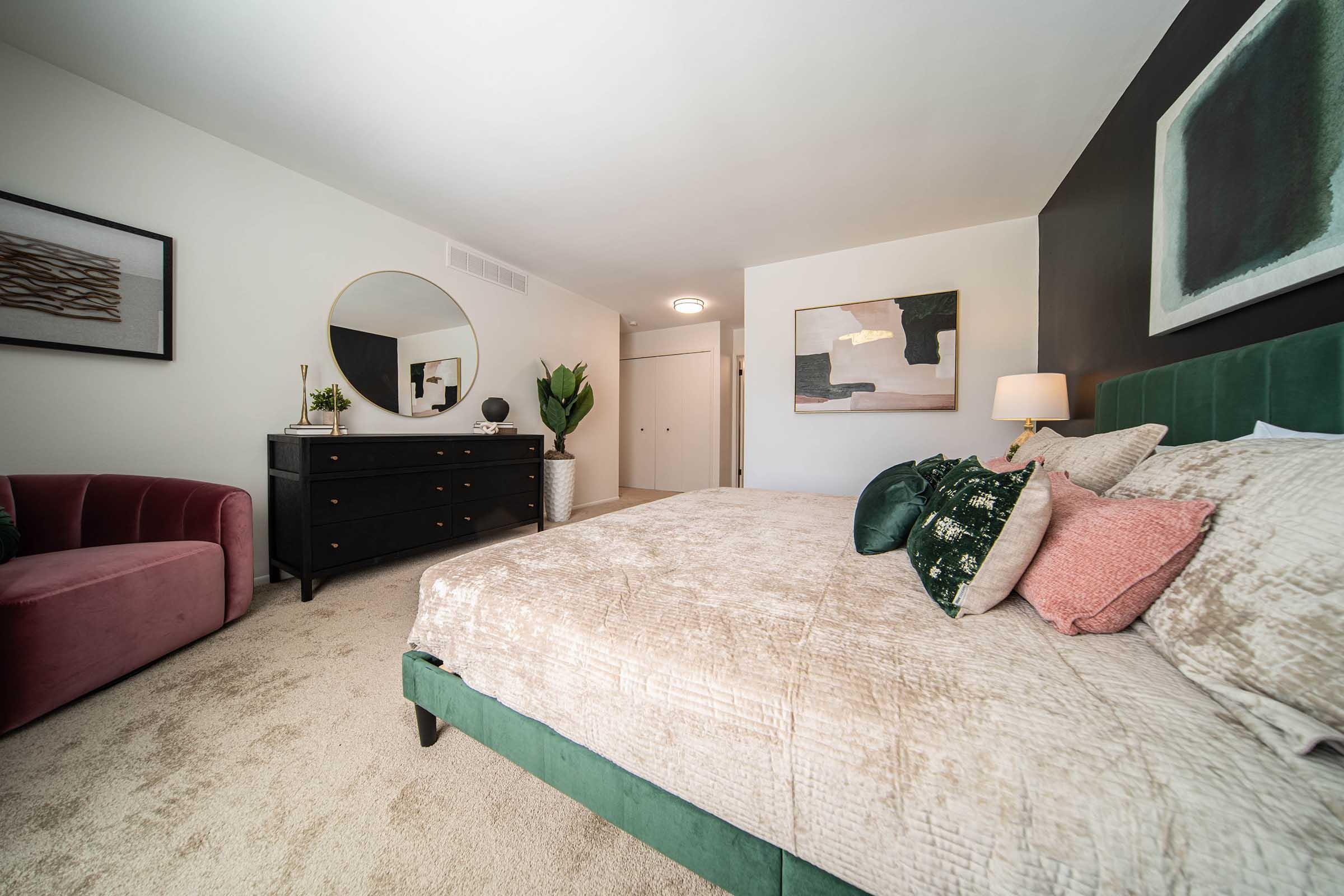 A cozy bedroom featuring a green upholstered bed with decorative pillows, a round mirror on the wall, a black dresser, a pink accent chair, a potted plant, and abstract wall art. The room is softly lit, creating a warm and inviting atmosphere.