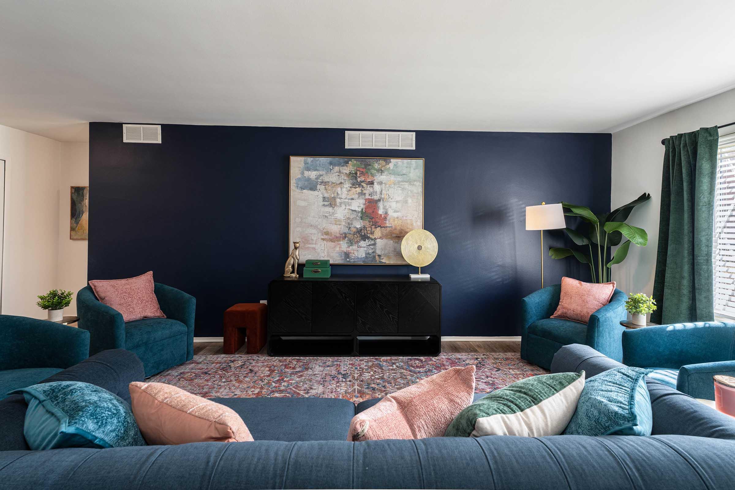 A modern living room featuring a deep blue accent wall, a large abstract painting, and a stylish black sideboard. Seating includes two teal armchairs and a sofa adorned with various colorful pillows. A floor lamp and potted plant add warmth to the space, complemented by a patterned area rug.