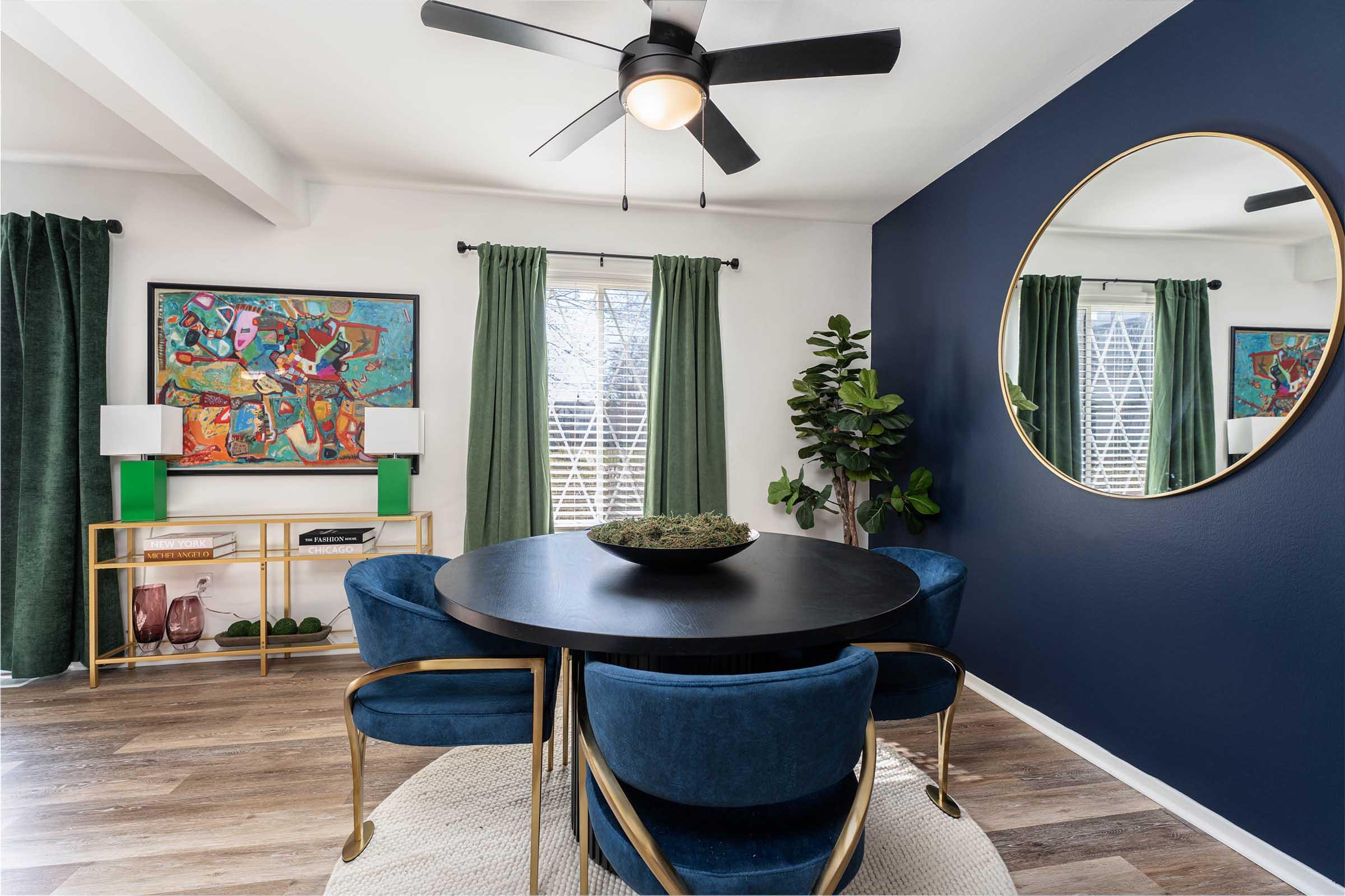 A modern dining area featuring a round black table surrounded by blue velvet chairs. There's a green plant in the corner, a decorative round mirror on the blue wall, and colorful artwork framed on the opposite wall. Natural light streams through the windows, with green curtains enhancing the decor.