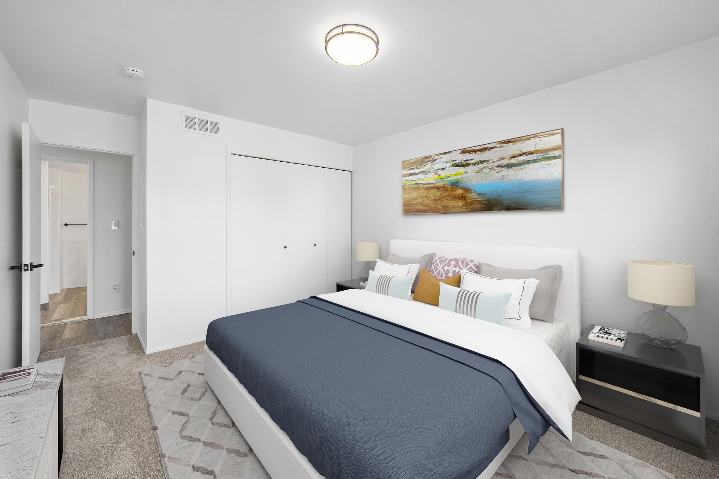 A modern bedroom featuring a neatly made bed with a blue blanket and accented pillows. There is a large piece of abstract wall art above the bed, beige carpet, and two nightstands with lamps. A closet is visible on the right, and a doorway leads to another room on the left, all set in a bright, neutral color palette.