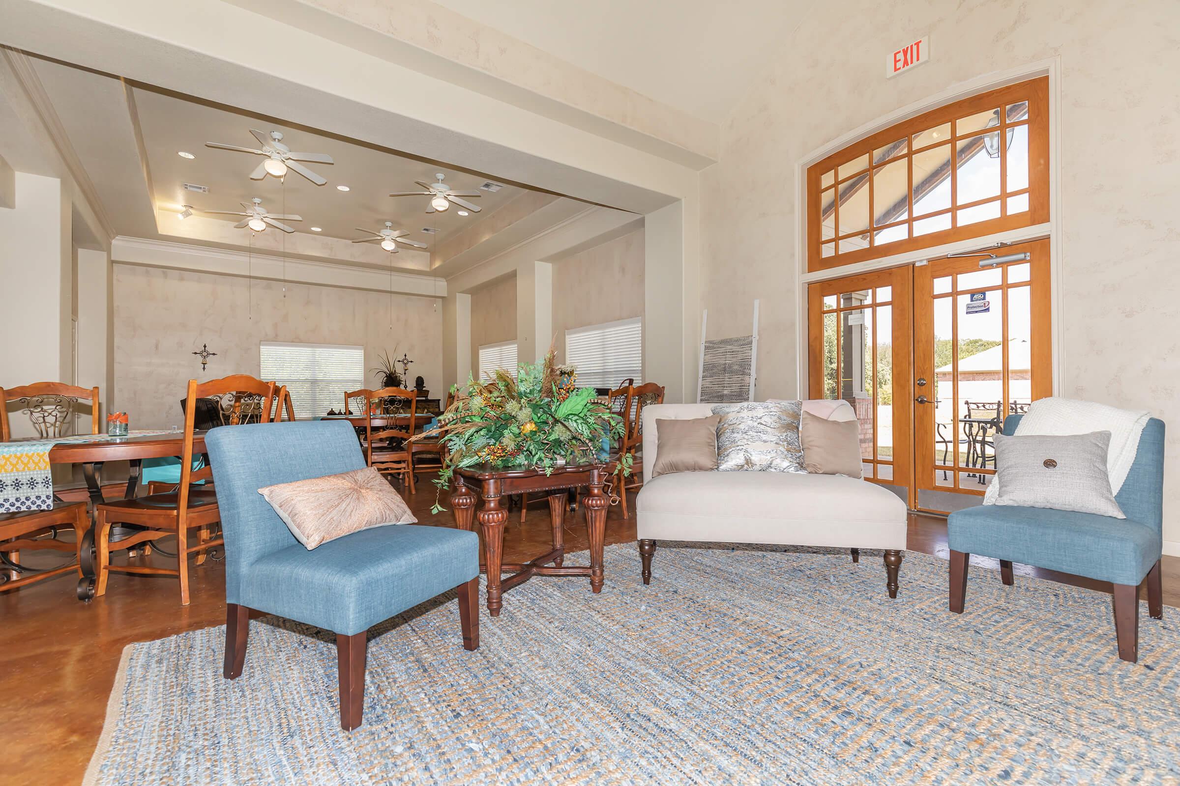 A cozy interior space featuring a mix of furniture. In the foreground, there are two blue chairs and a beige chair with decorative pillows, alongside a coffee table with green plants. The background shows a dining area with wooden tables and chairs, and large windows allowing natural light to fill the room.