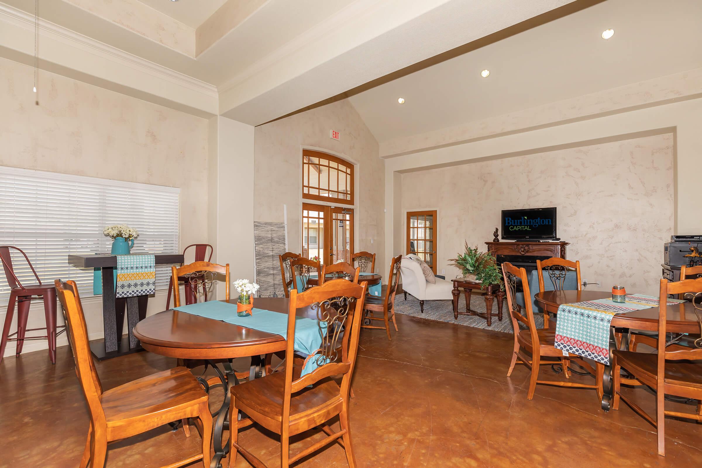 A spacious, well-lit social area featuring several wooden tables with chairs, a small blue table with flowers, and a cozy seating arrangement. Large windows provide natural light, and a TV can be seen in the background. The decor blends modern and traditional elements.