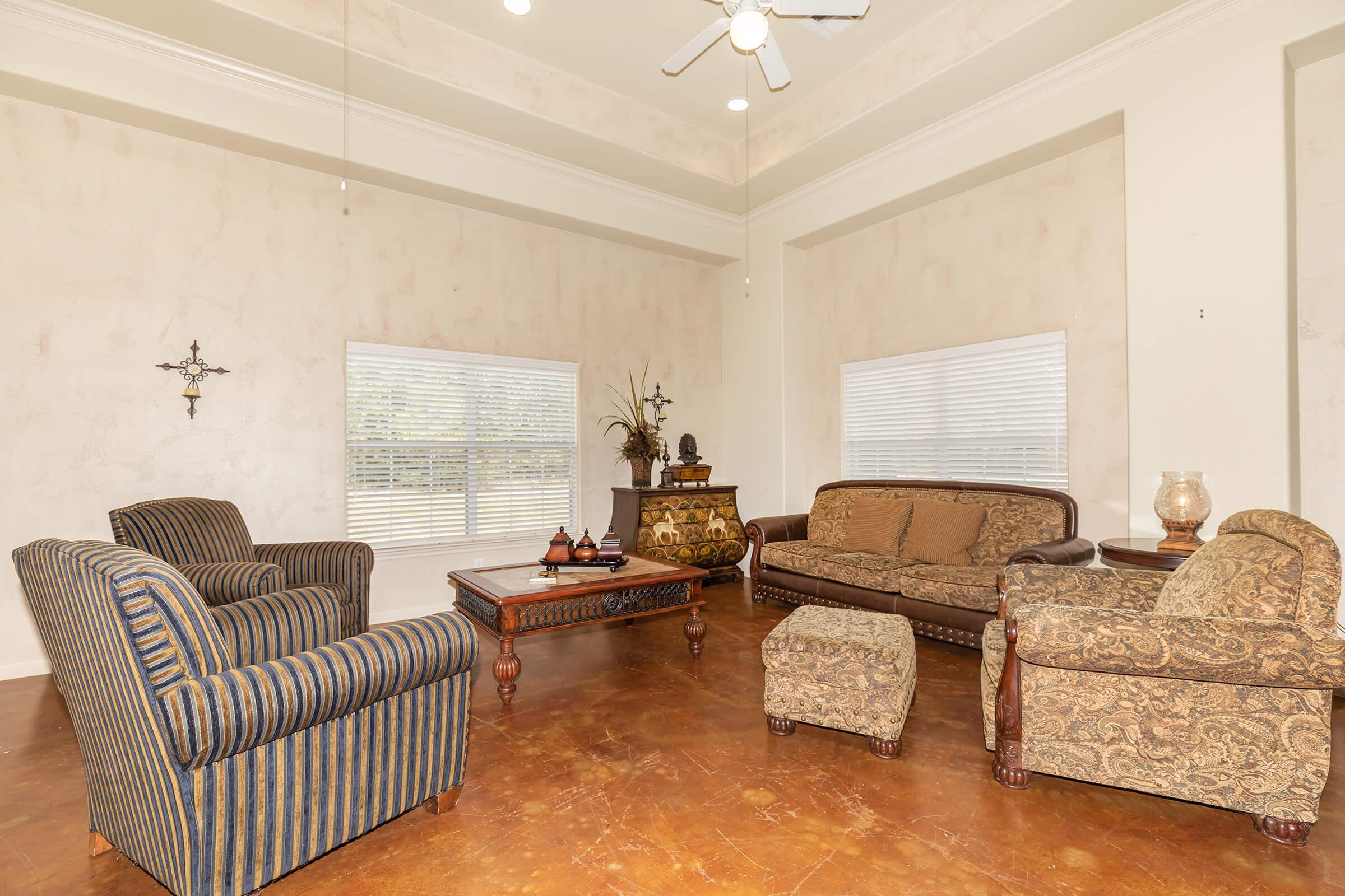 A cozy living room featuring a patterned couch and armchairs, a decorative coffee table, and a small footstool. Large windows let in natural light, and the walls have a subtle texture. The space is adorned with plants and a wall decoration, creating a warm and inviting atmosphere.