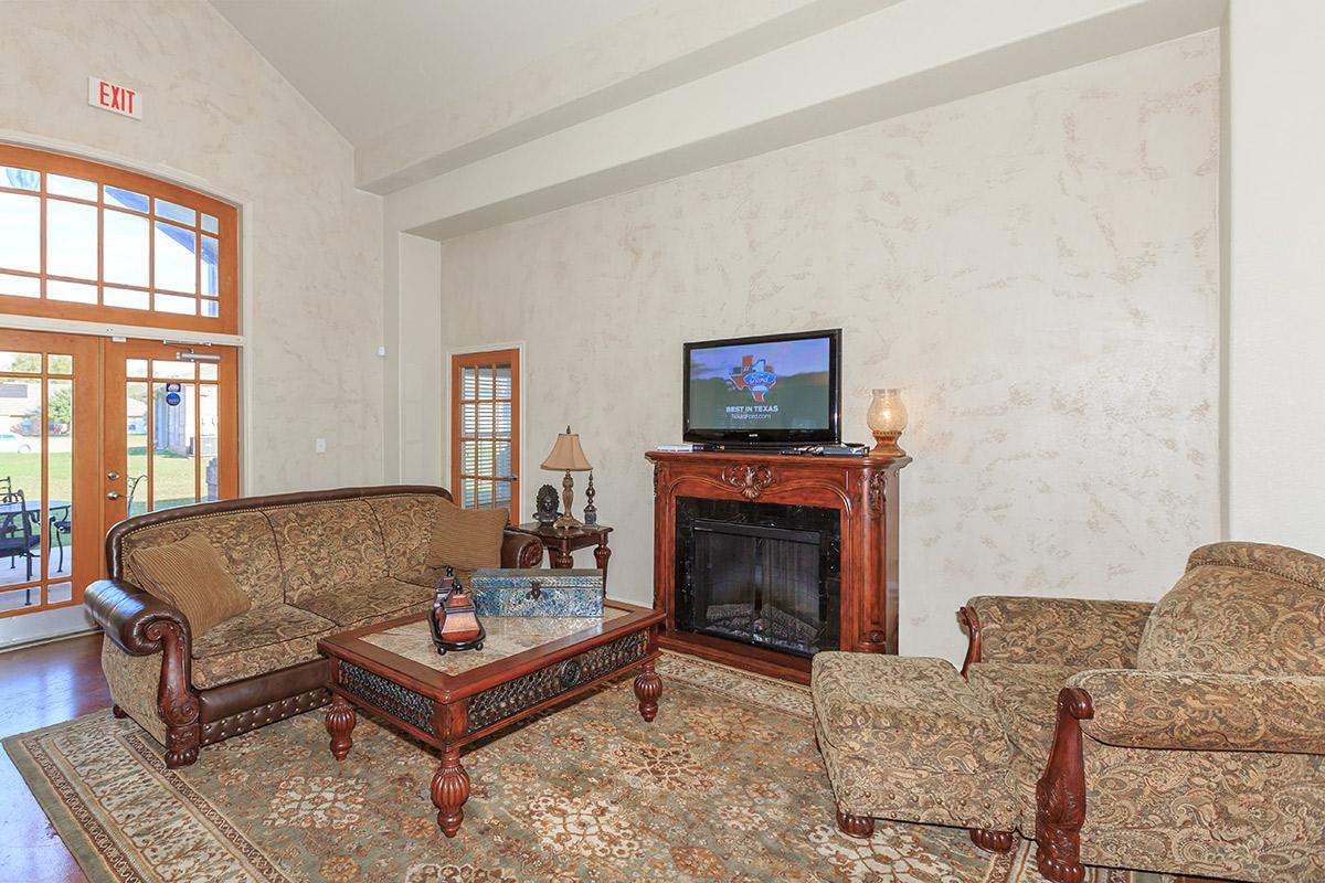A cozy living room featuring a large sofa and an armchair, arranged around a decorative coffee table. A wooden fireplace is flanked by a lamp and a television mounted on the wall. Light-colored walls and a large window provide a bright atmosphere, with a patterned rug on the wooden floor.